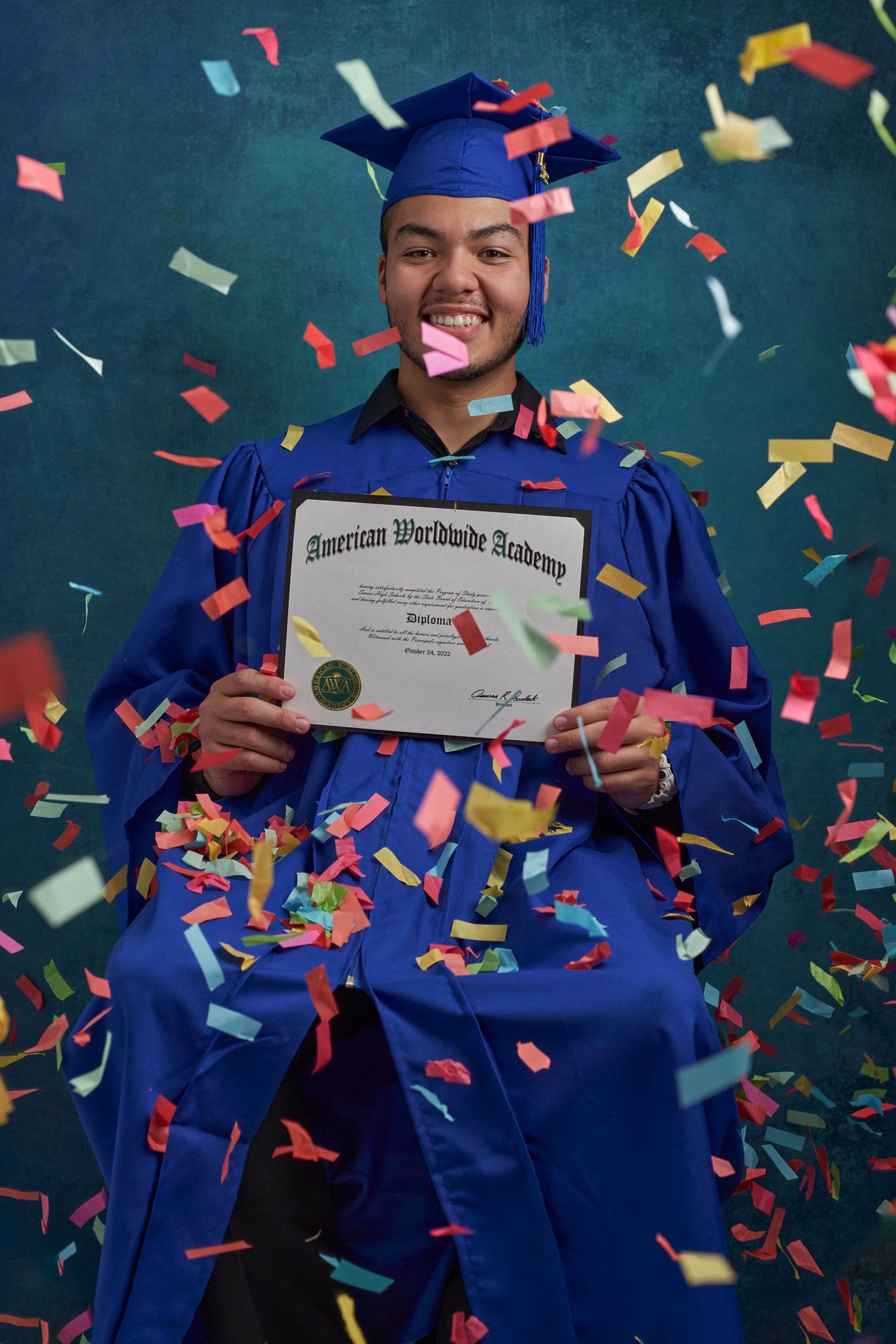 Smiling graduate in blue cap and gown holds diploma surrounded by colorful falling confetti.
