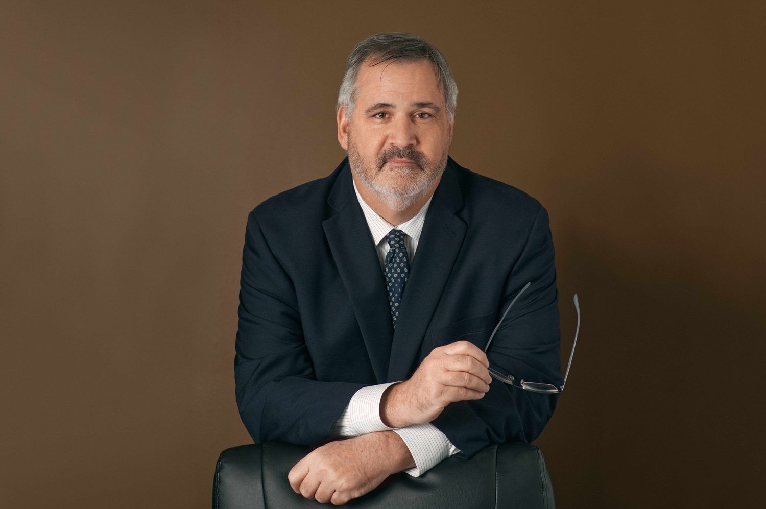 Professional man in dark blazer holding glasses while seated, posing confidently against warm brown background.