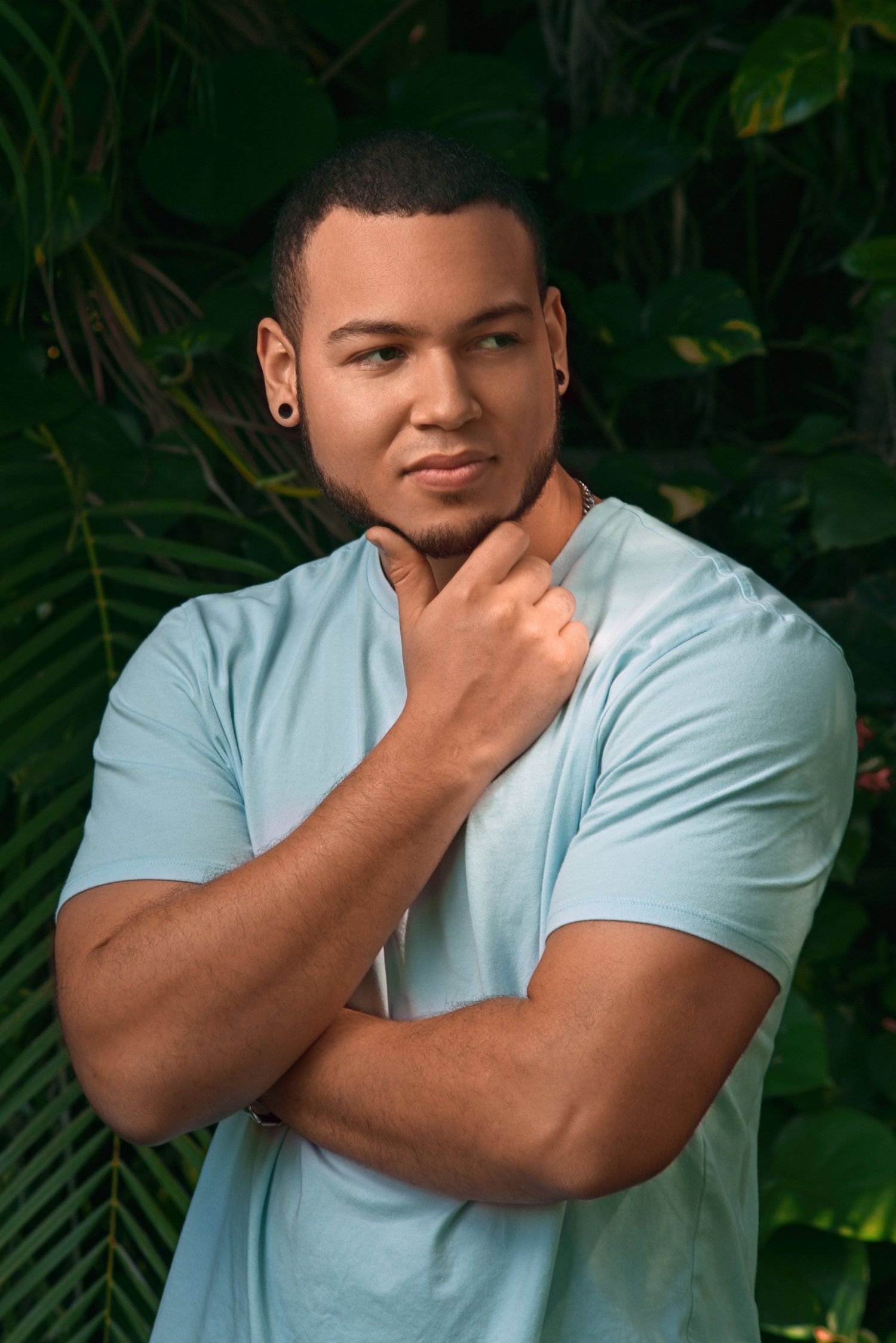 Muscular young man in light blue tee poses thoughtfully against tropical greenery.