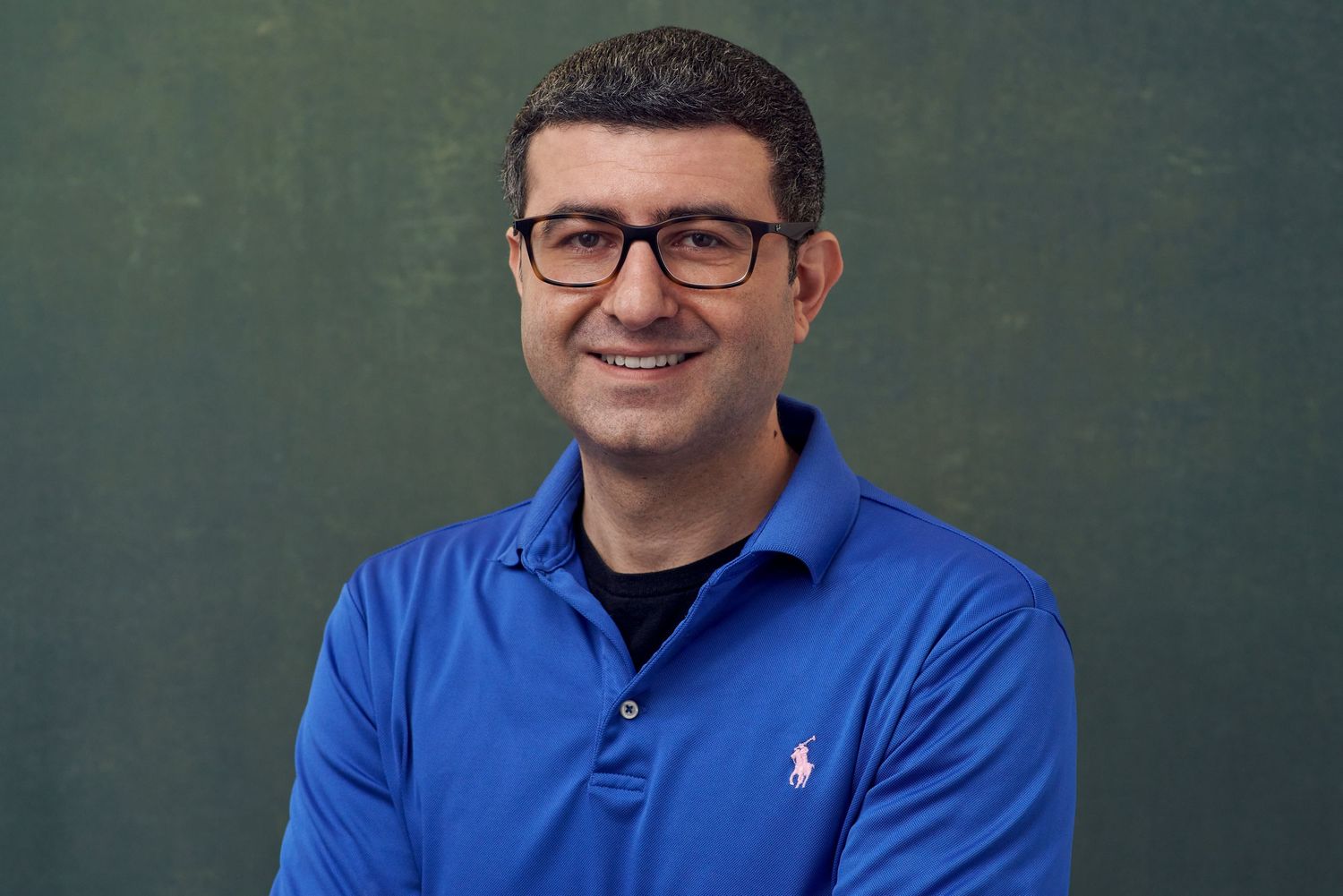 Smiling man in blue polo shirt wearing glasses poses against green background.