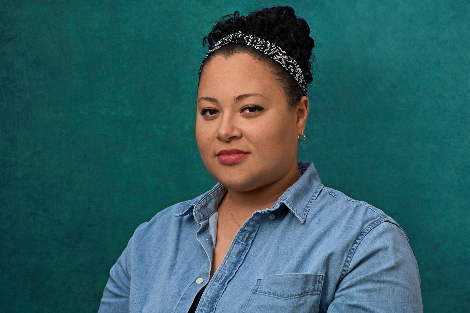 Woman with natural updo and bandana headband poses in denim shirt against teal background.