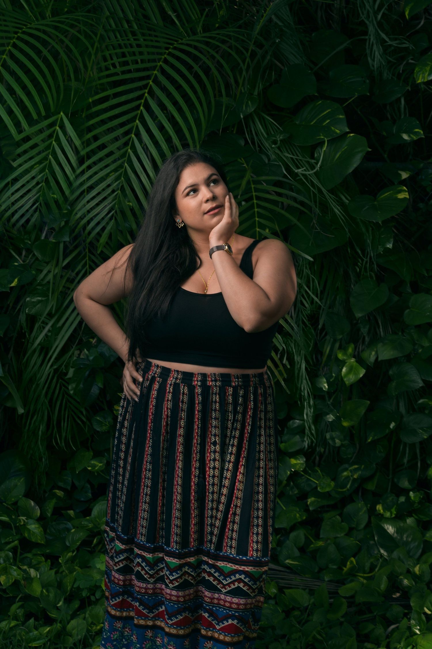 Woman in black crop top and patterned skirt poses against lush tropical foliage.