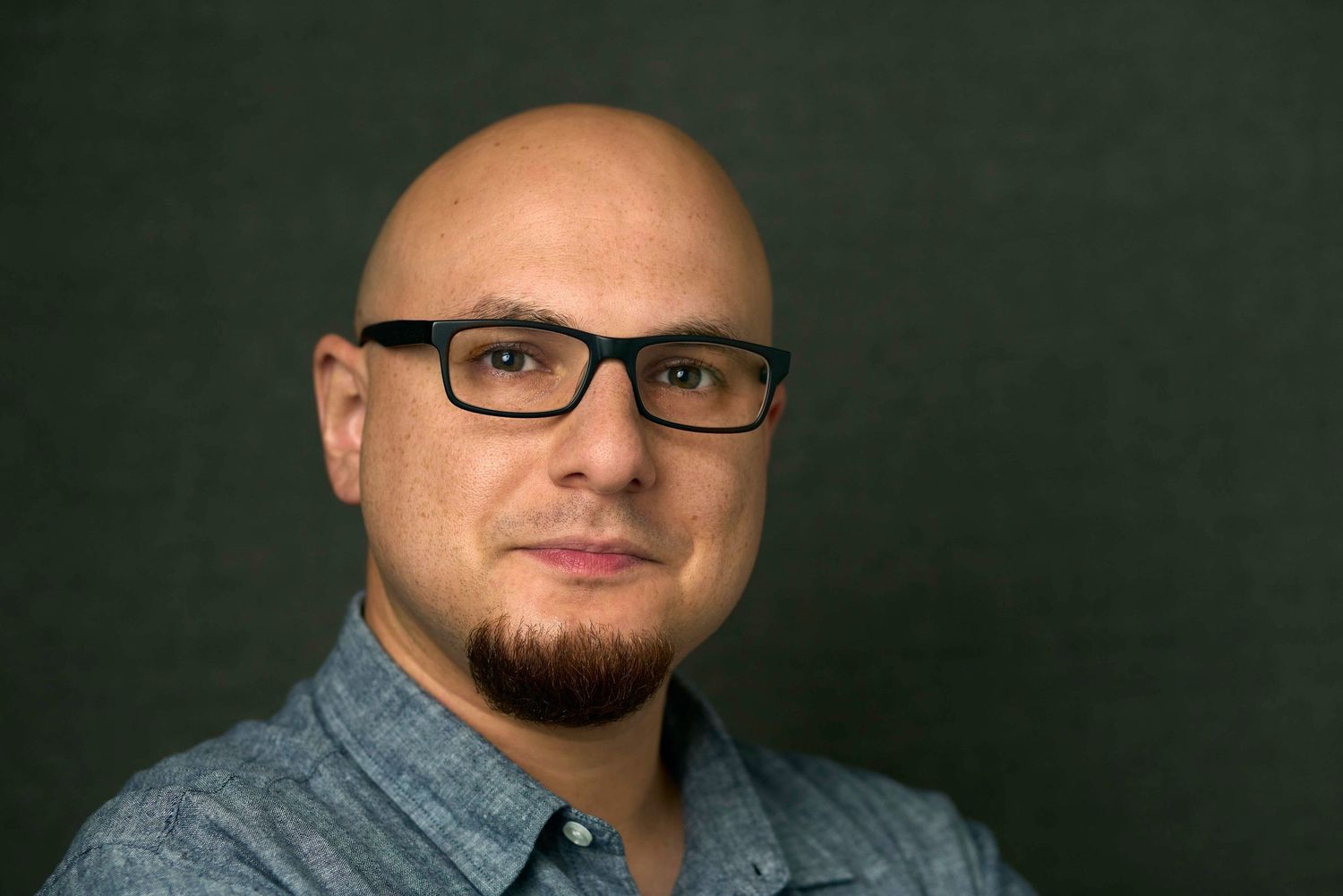 Bald man with black glasses and goatee wearing a blue shirt, posing against a dark background.