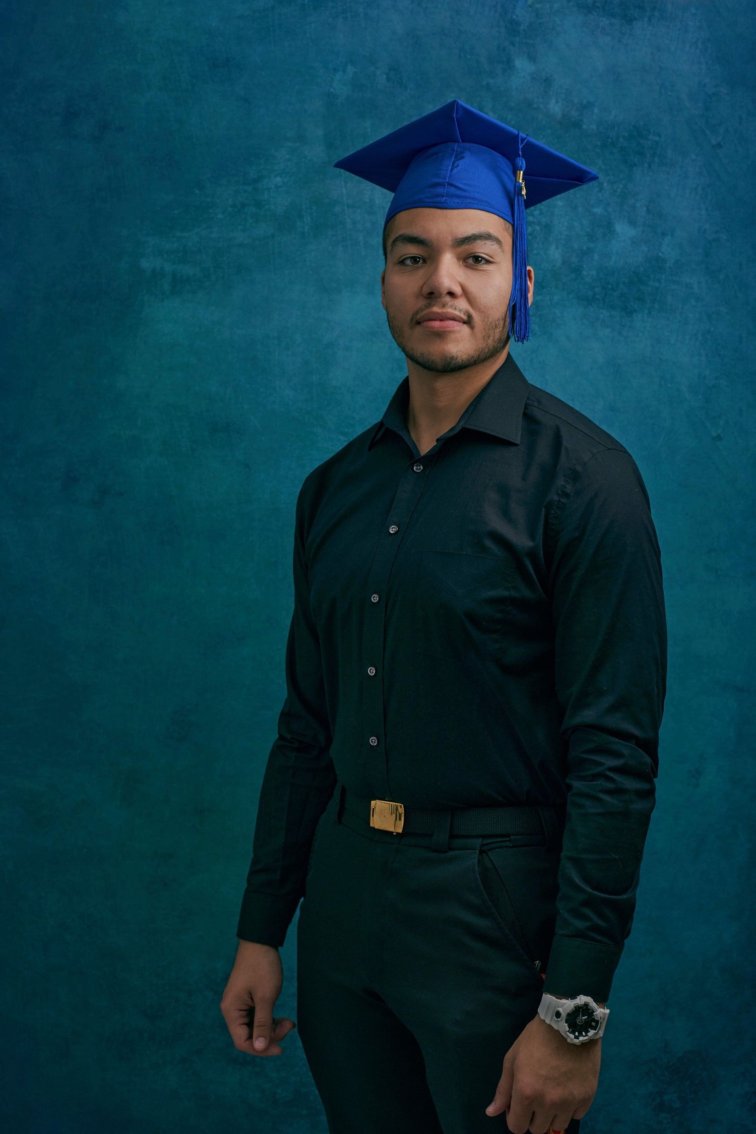 Young man in black shirt and blue graduation cap poses for portrait photo against teal background.