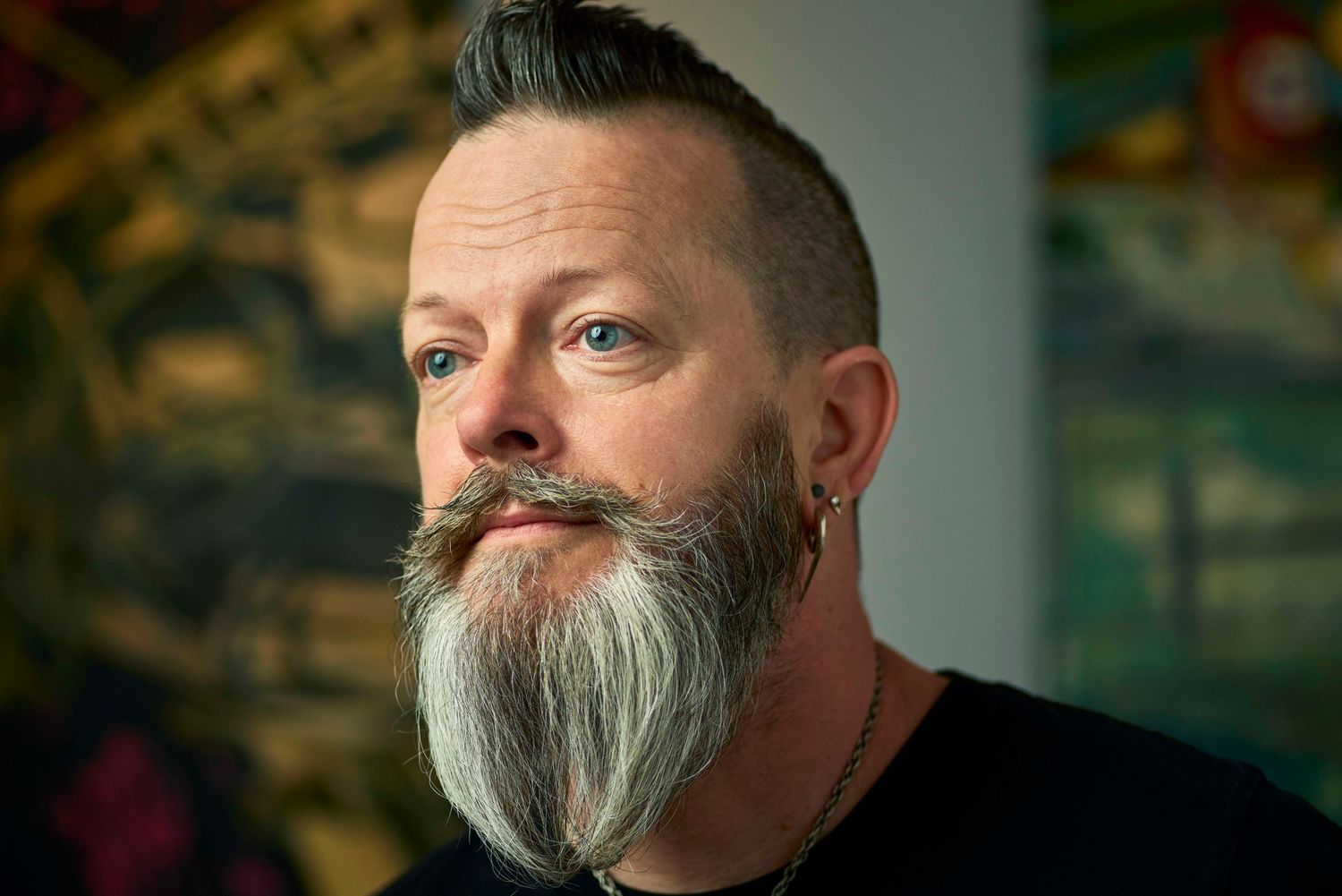 Close-up portrait of man with distinctive gray-streaked beard and mohawk hairstyle against a vibrant painted backdrop.