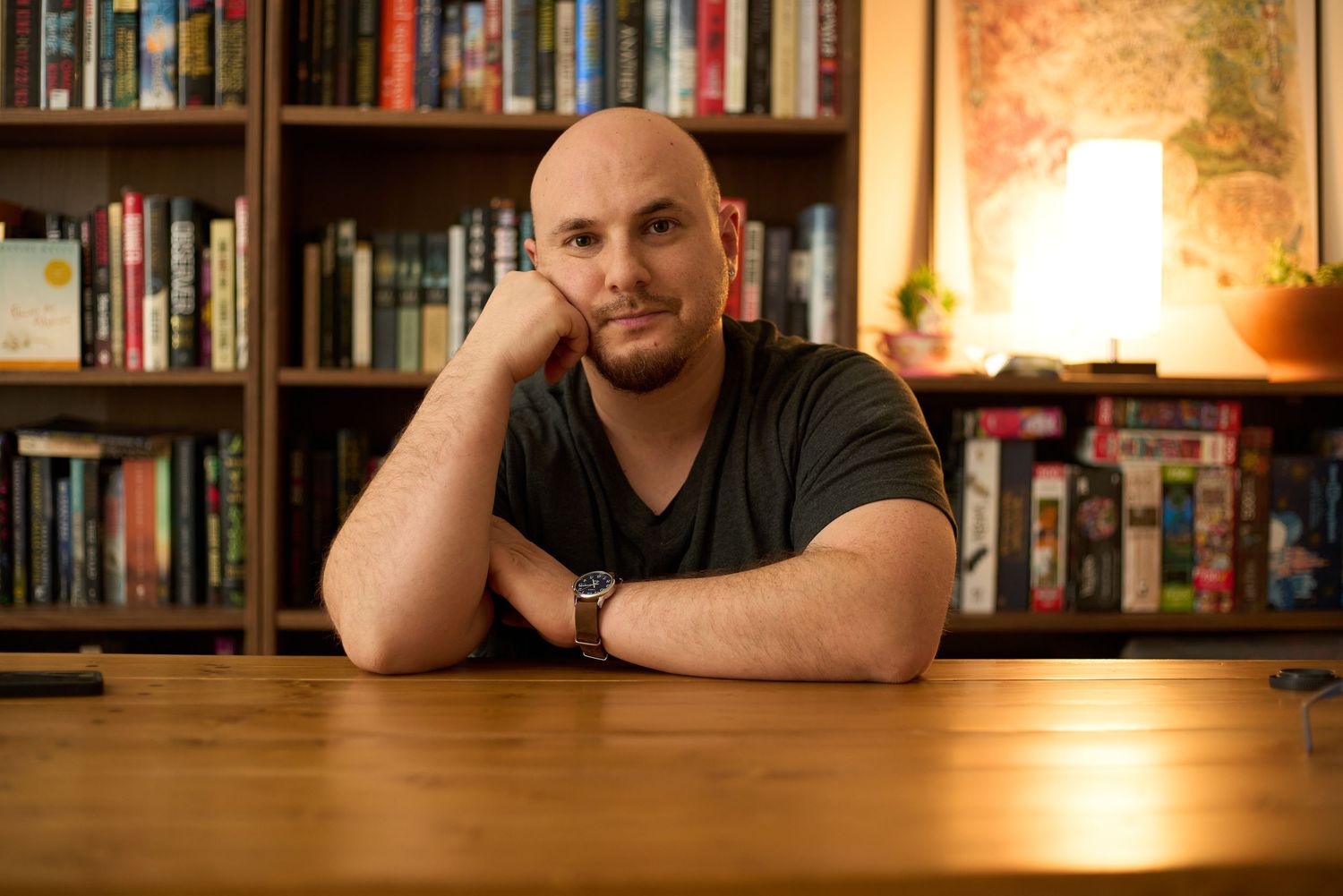 Man with beard resting chin on hand at wooden table with bookshelf and warm lamp in background.
