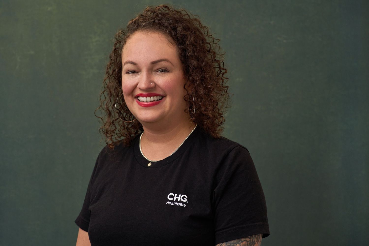Smiling woman with curly hair wearing black CHG Healthcare shirt against dark green background.