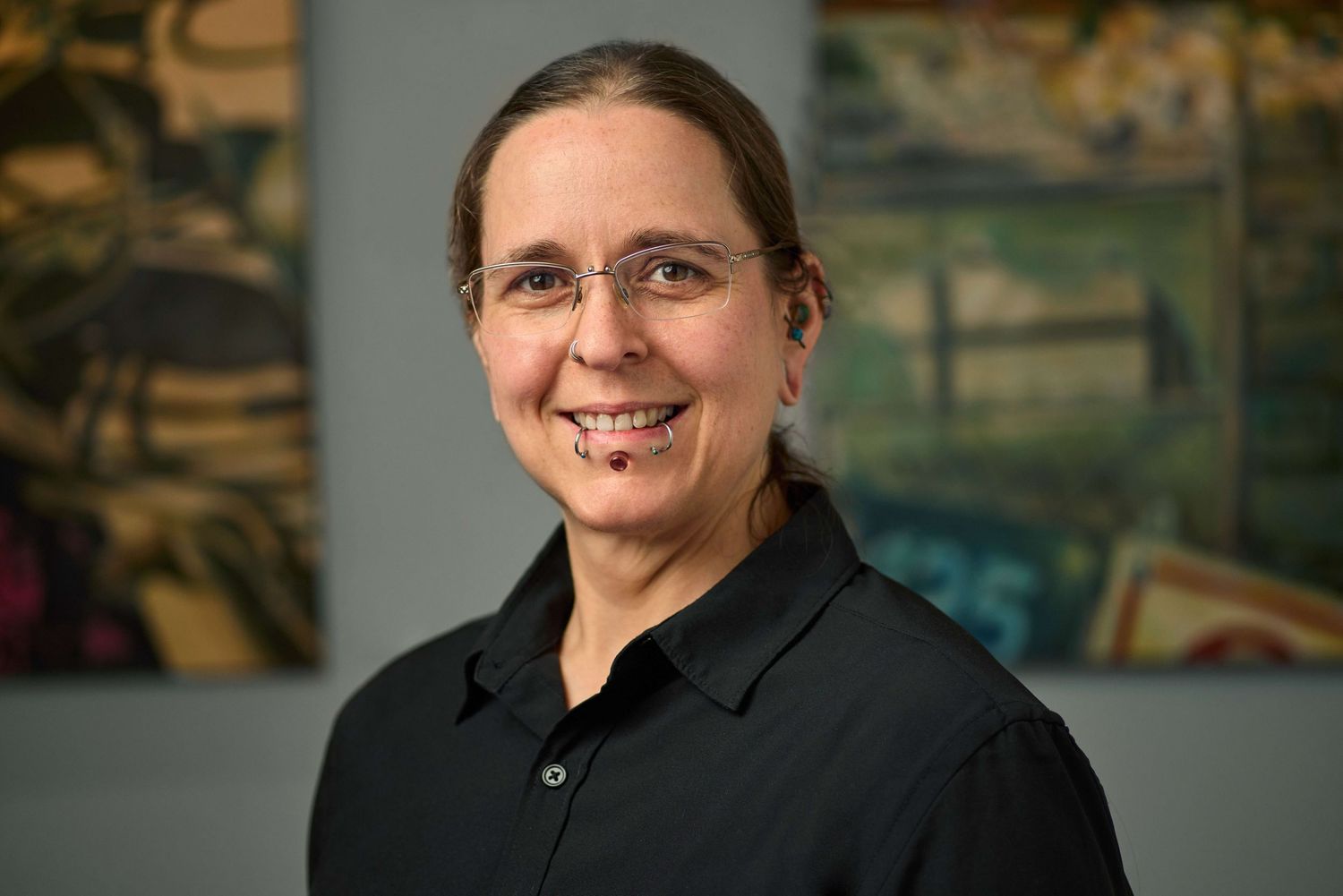 Smiling woman with glasses in black shirt poses against vibrant painted artwork background.