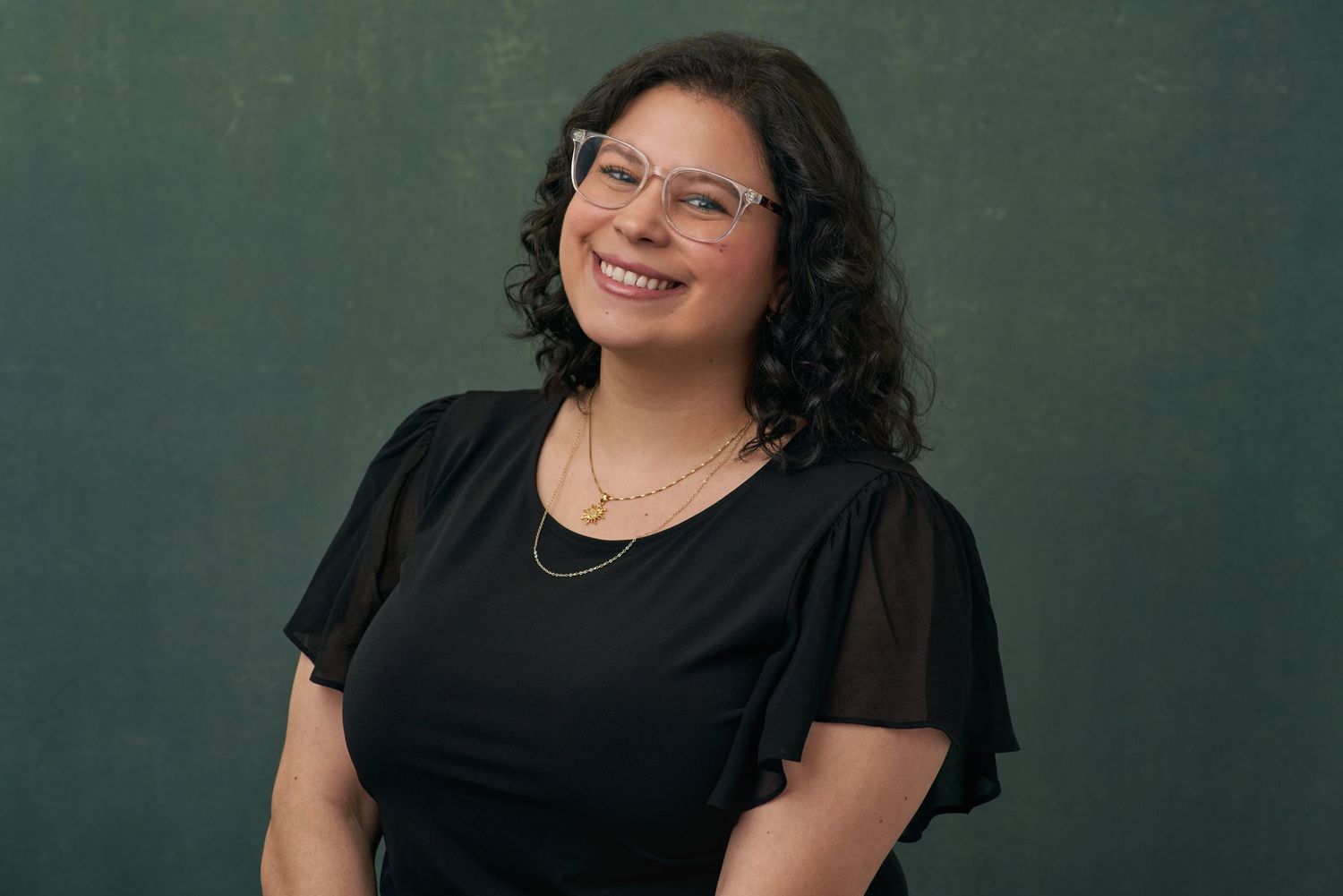 Smiling woman with curly hair and glasses wearing black top against green backdrop.