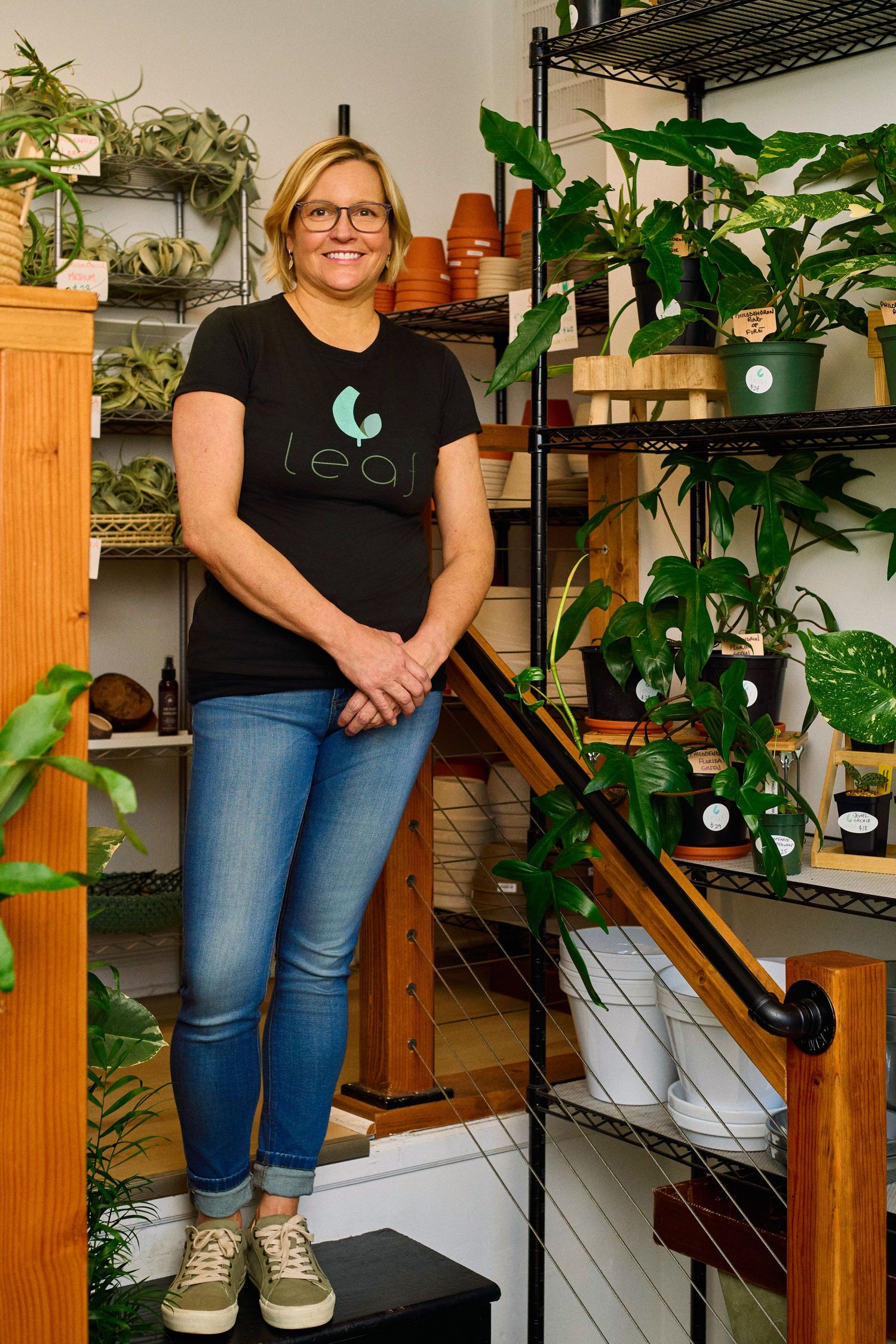 Smiling woman in Leaf branded black t-shirt and jeans stands among shelves of lush houseplants in a plant shop.