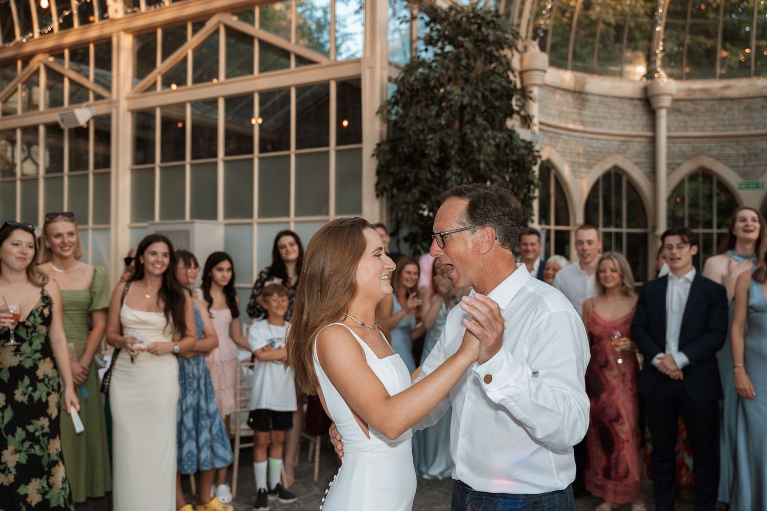 A couple shares their first dance at a wedding reception while guests look on in a beautiful greenhouse setting.