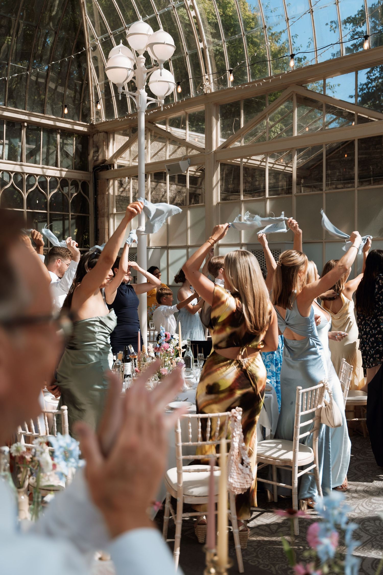 Wedding guests cheer and wave napkins in celebration at an indoor reception venue with glass ceiling and ornate lighting.