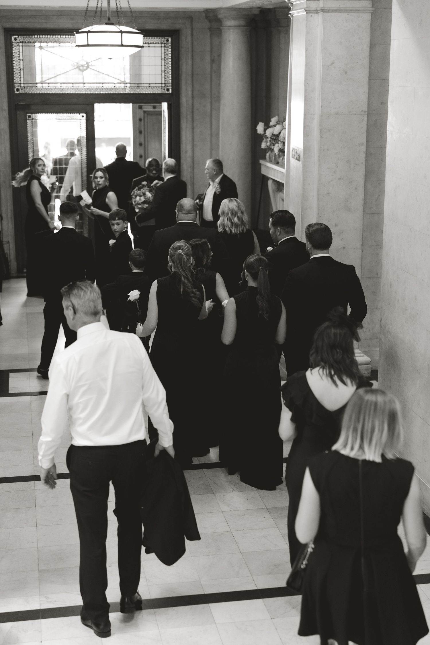 Black and white photo of formal event attendees gathering in an elegant hallway with columns and chandeliers.