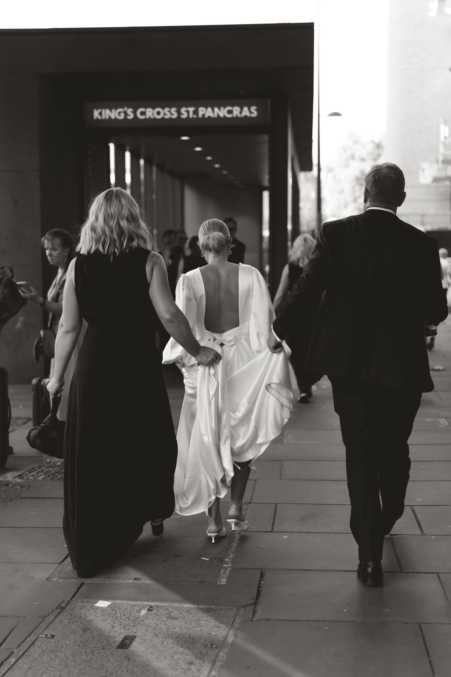 A wedding party walks through King's Cross St Pancras station with the bride holding up her white dress.