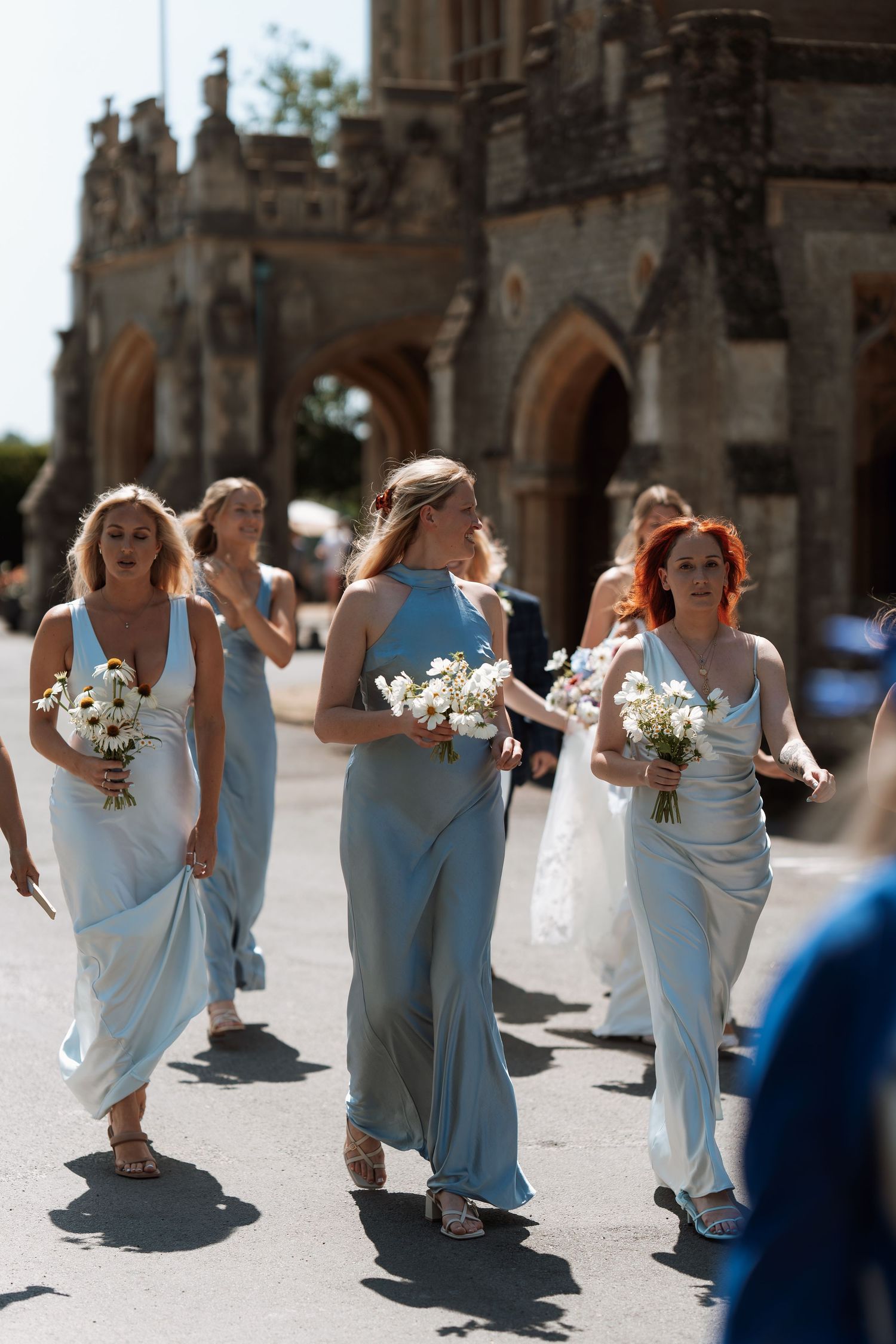 Wedding party dressed in elegant light blue gowns walks along stone building courtyard holding white floral bouquets.