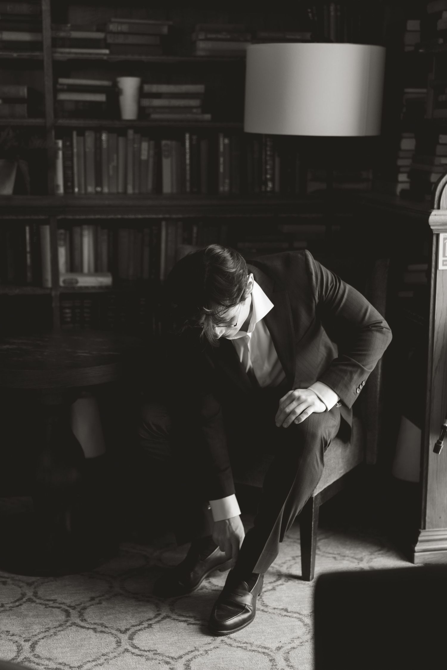 Black and white photo showing a person adjusting their shoe in a cozy library setting with bookshelves and a lamp.