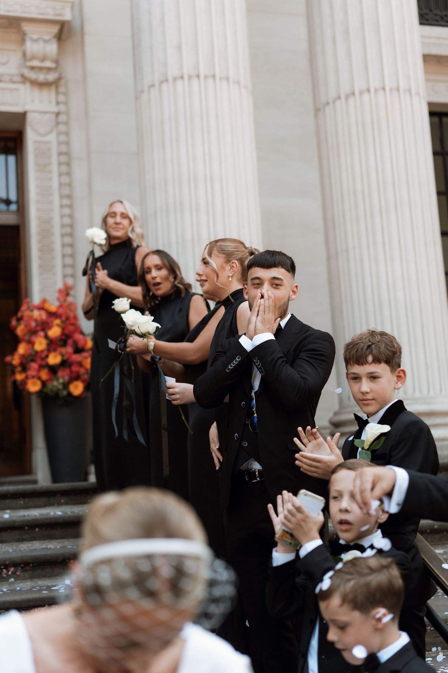 Wedding guests in formal attire react emotionally on elegant steps with white columns and floral decorations.