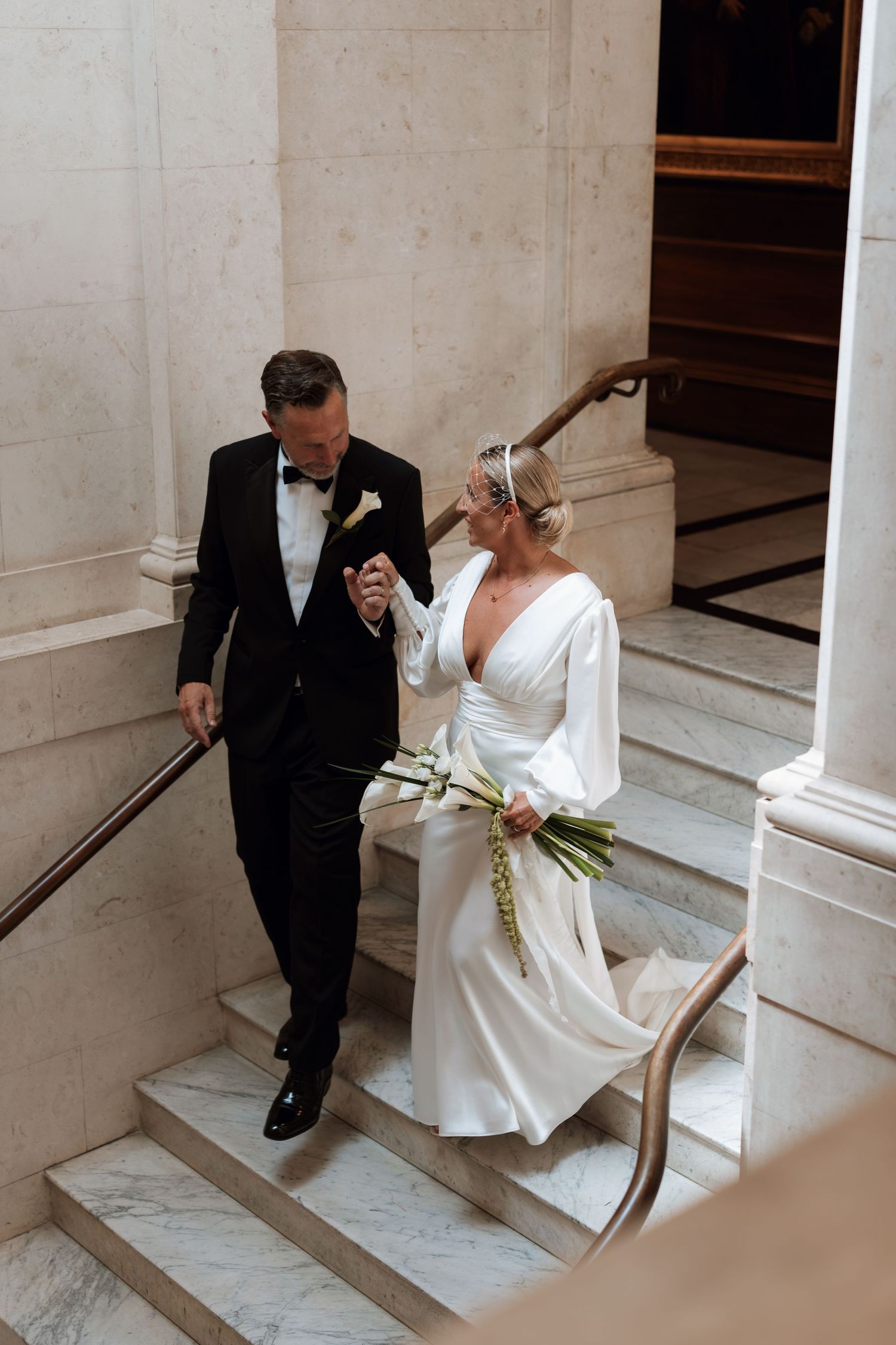 Elegant couple descending marble stairs, bride wearing white dress with long sleeves.