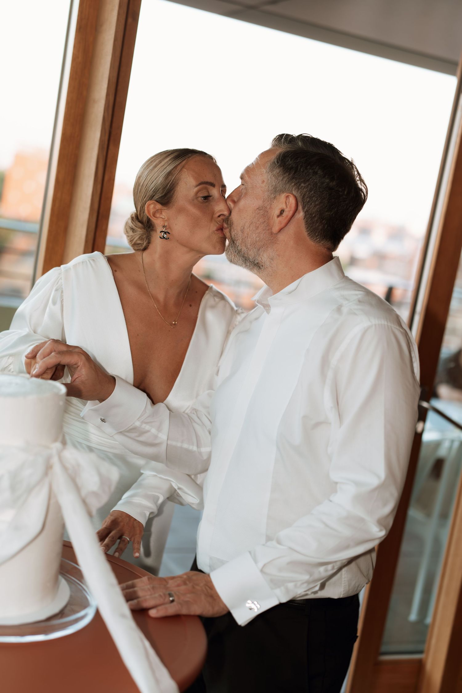 A couple shares an intimate moment in formal attire by a window.