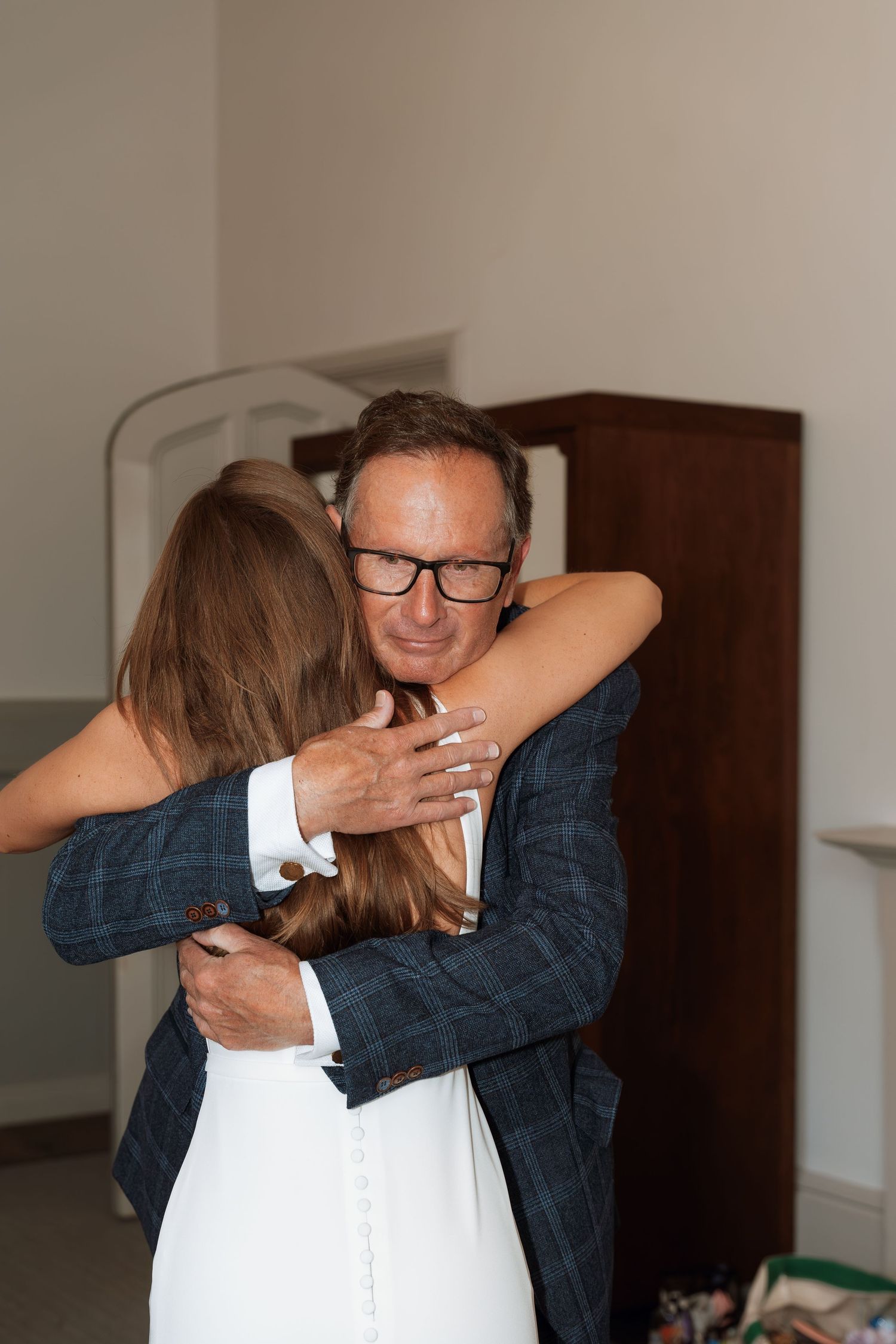 A loving embrace between two people at an indoor gathering, with one wearing a white dress and another in a navy blazer.