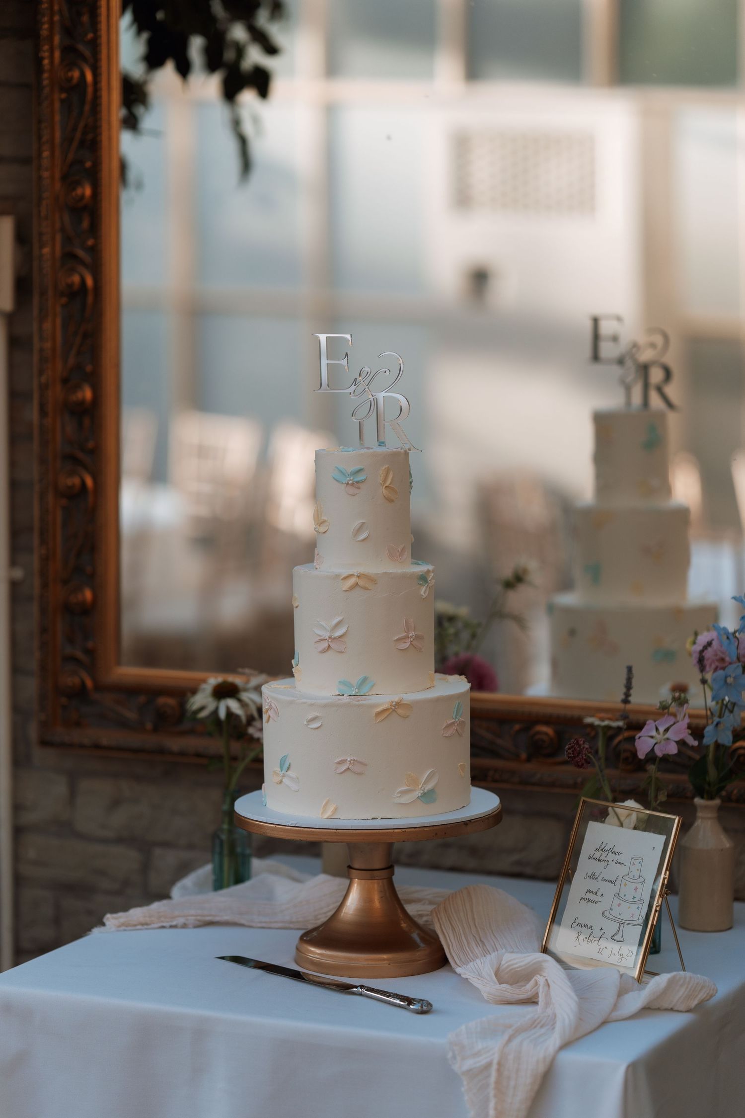 Elegant three-tier white wedding cake with delicate leaf detailing sits on gold stand beside framed sign at reception table.