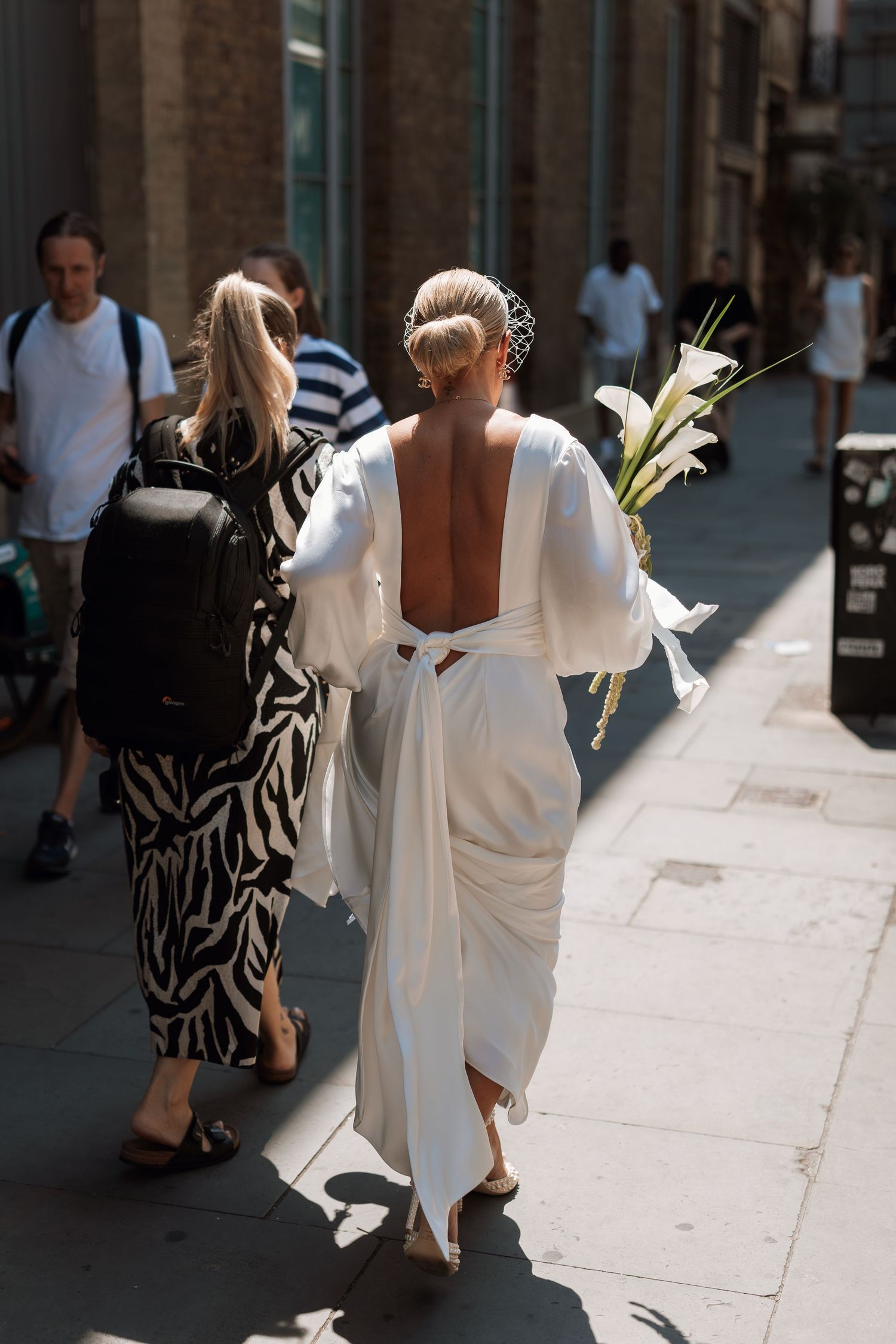 People walking on a city sidewalk with one wearing a flowing white backless dress and carrying flowers on a sunny day.