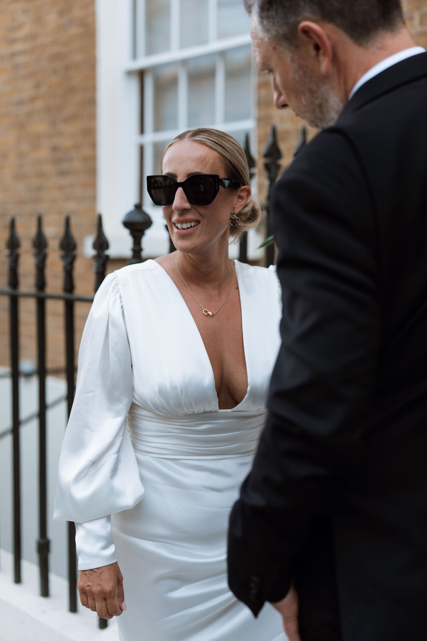 Elegant bride in white dress with sunglasses smiling outside a brick building with iron railings.