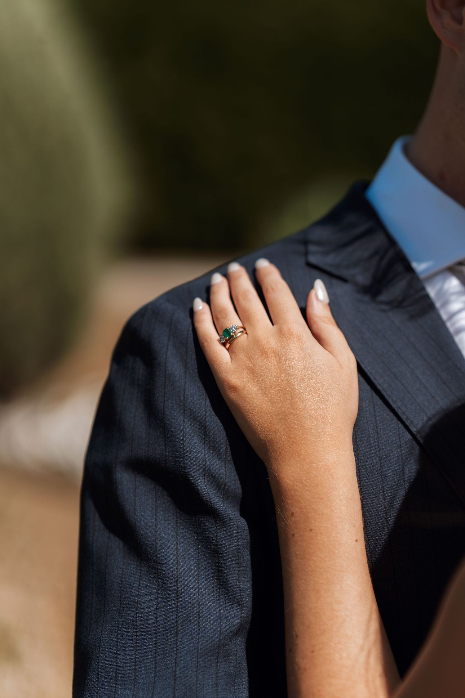 A hand wearing a green gemstone ring rests on a navy pinstriped suit jacket shoulder showing elegant wedding details.