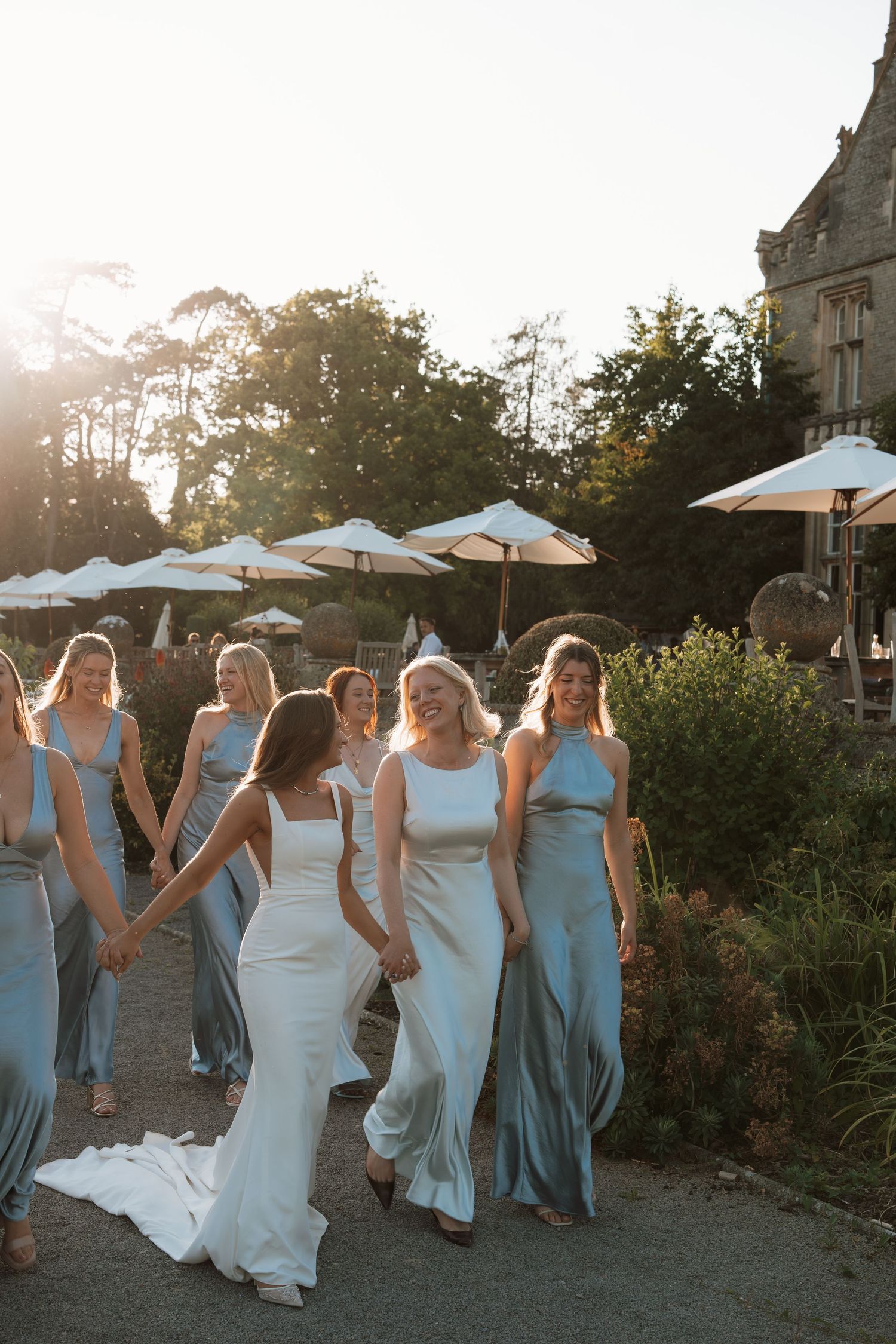 A group in coordinating light blue bridesmaid dresses and white wedding gown walking together in sunset garden setting.