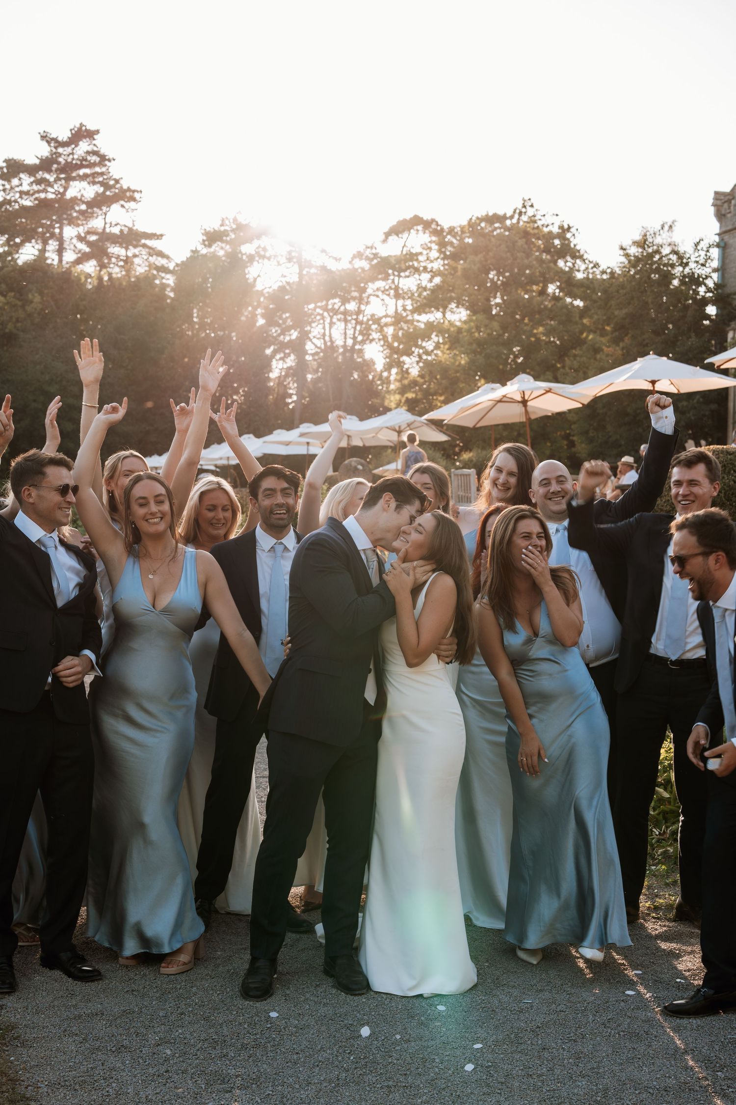 Wedding party celebrates outdoors at sunset with raised arms and joyful expressions.
