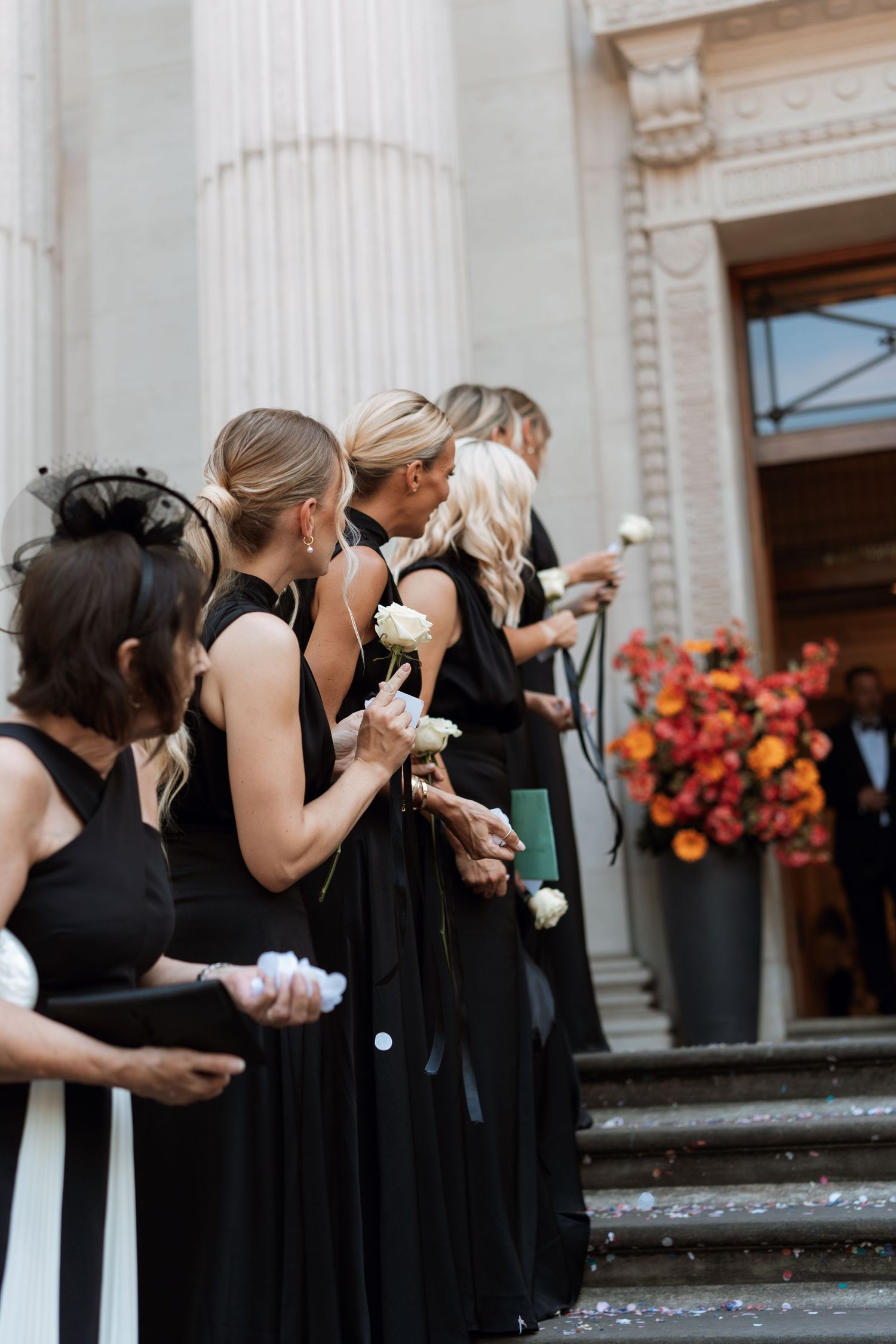 Bridesmaids in elegant black dresses stand in line on stone steps holding flowers near classical white columns.