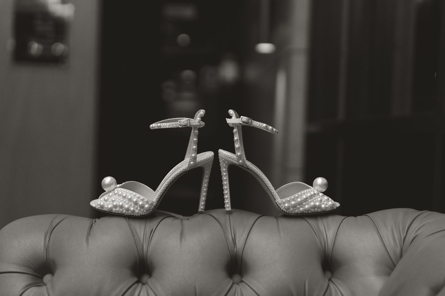 A pair of elegant silver wedding shoes positioned on a gray tufted leather couch in black and white photography.