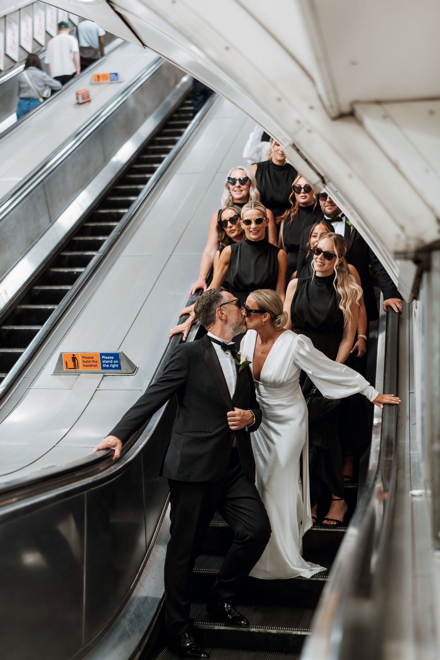 Wedding party in black and white attire riding an escalator with bridesmaids wearing sunglasses.