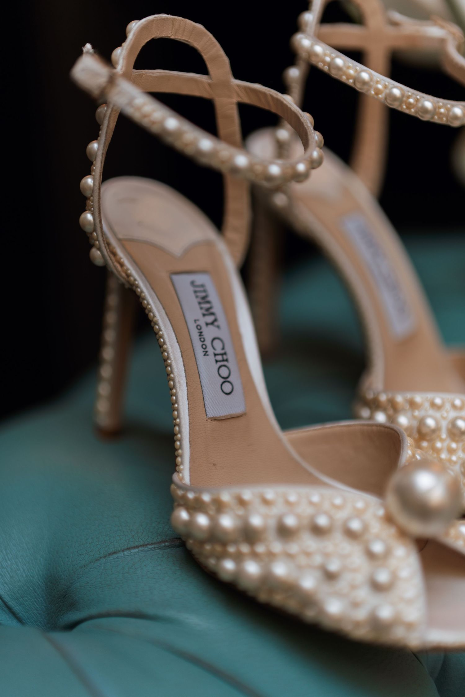 Elegant Jimmy Choo bridal shoes adorned with pearls and crystals sit on a teal surface.