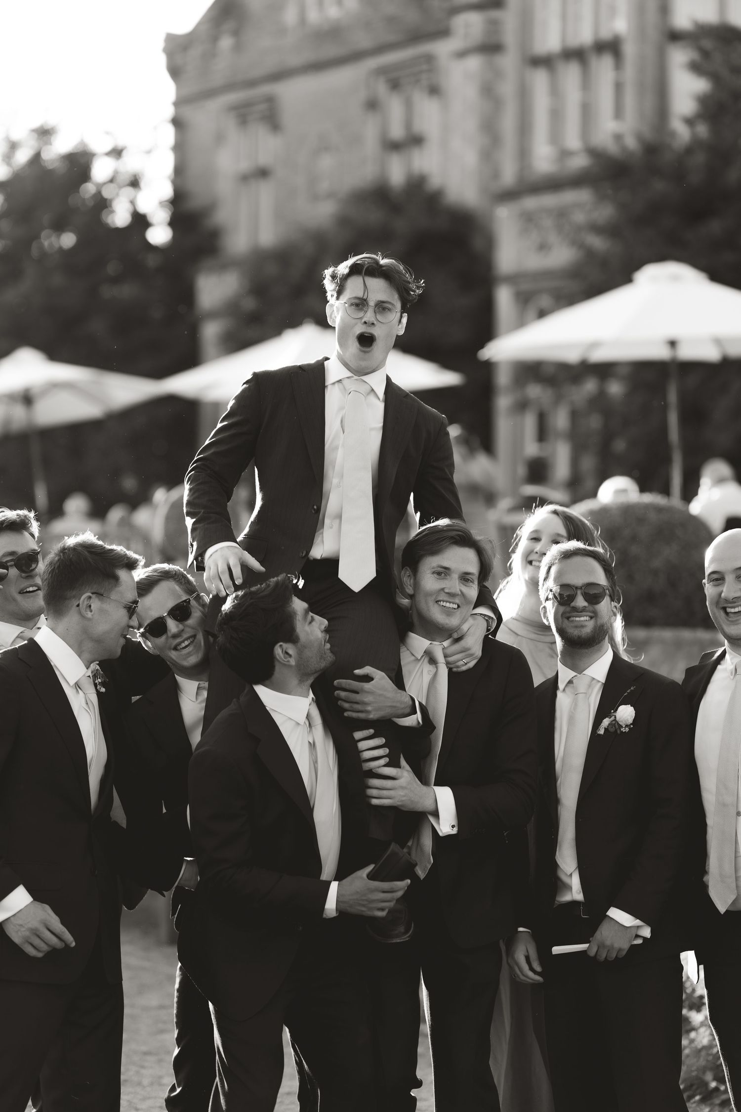 Wedding party lifts groomsman in black tuxedo and white tie during outdoor formal celebration.