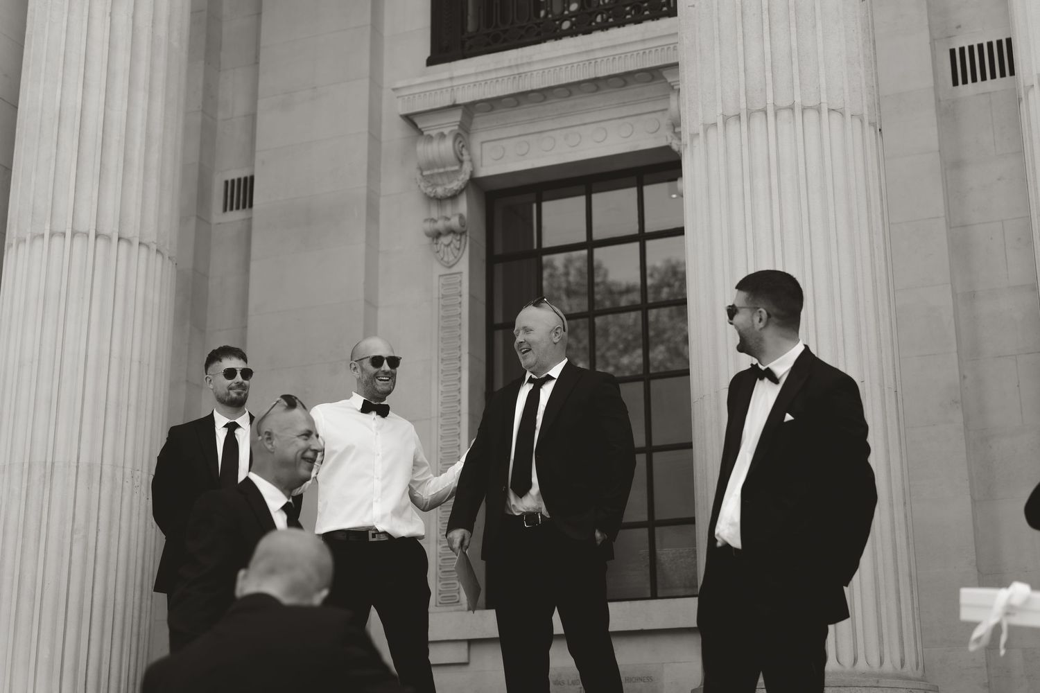 People in formal attire standing near white pillars and columns of a classical-style building exterior.