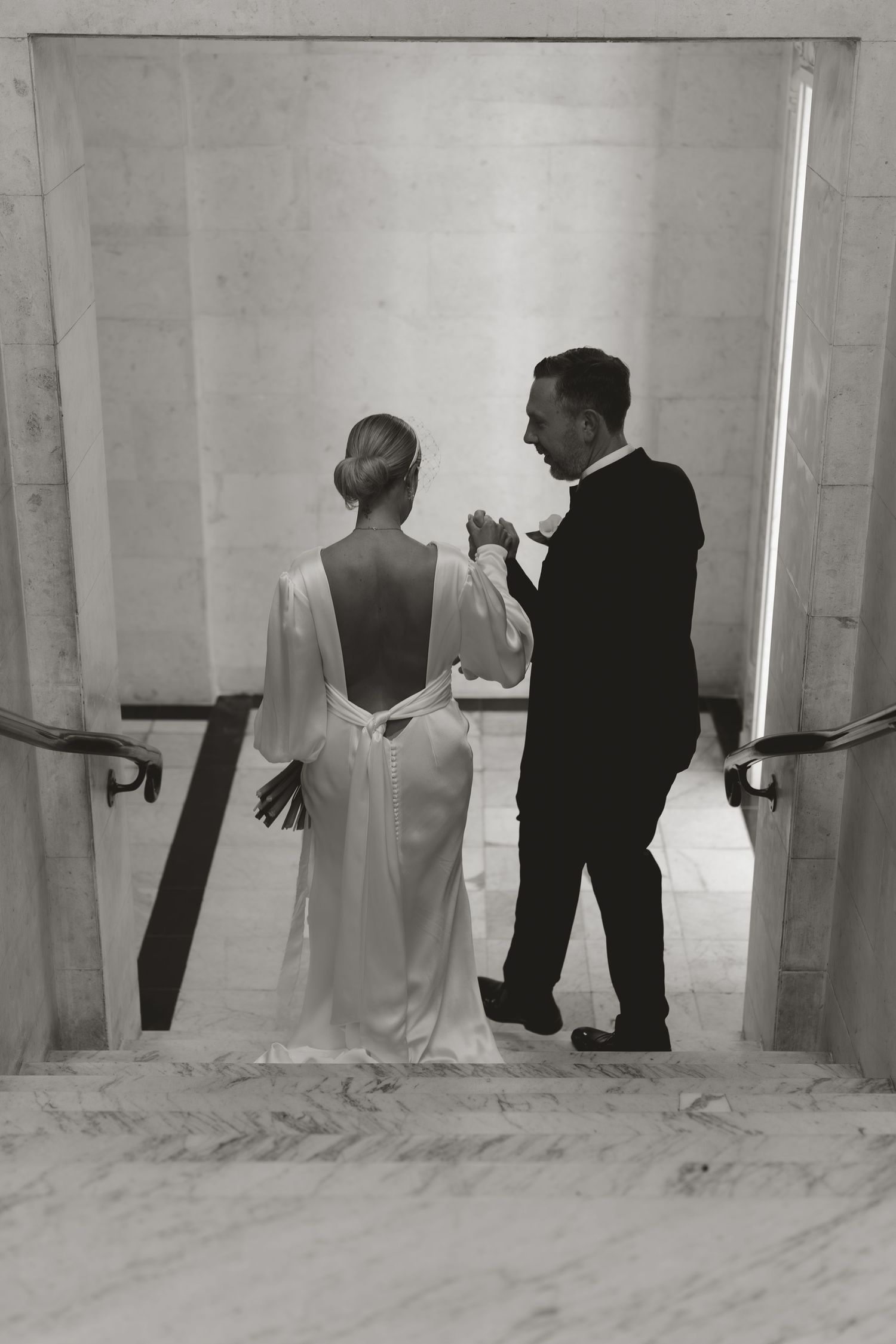 Black and white photo of a couple descending marble stairs in elegant formal attire.