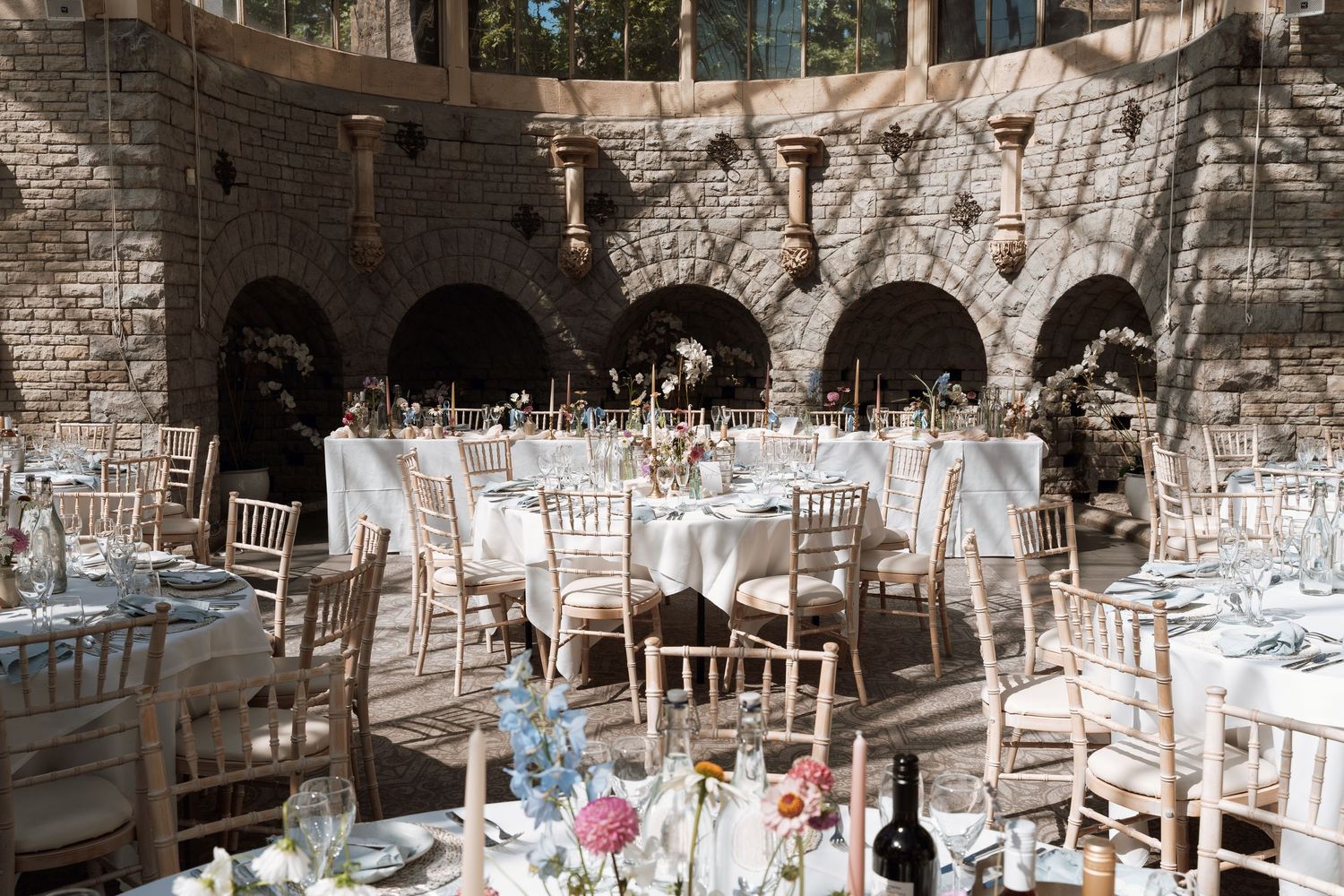 Elegant outdoor wedding reception setup with white chairs and tables in a stone arched courtyard setting.