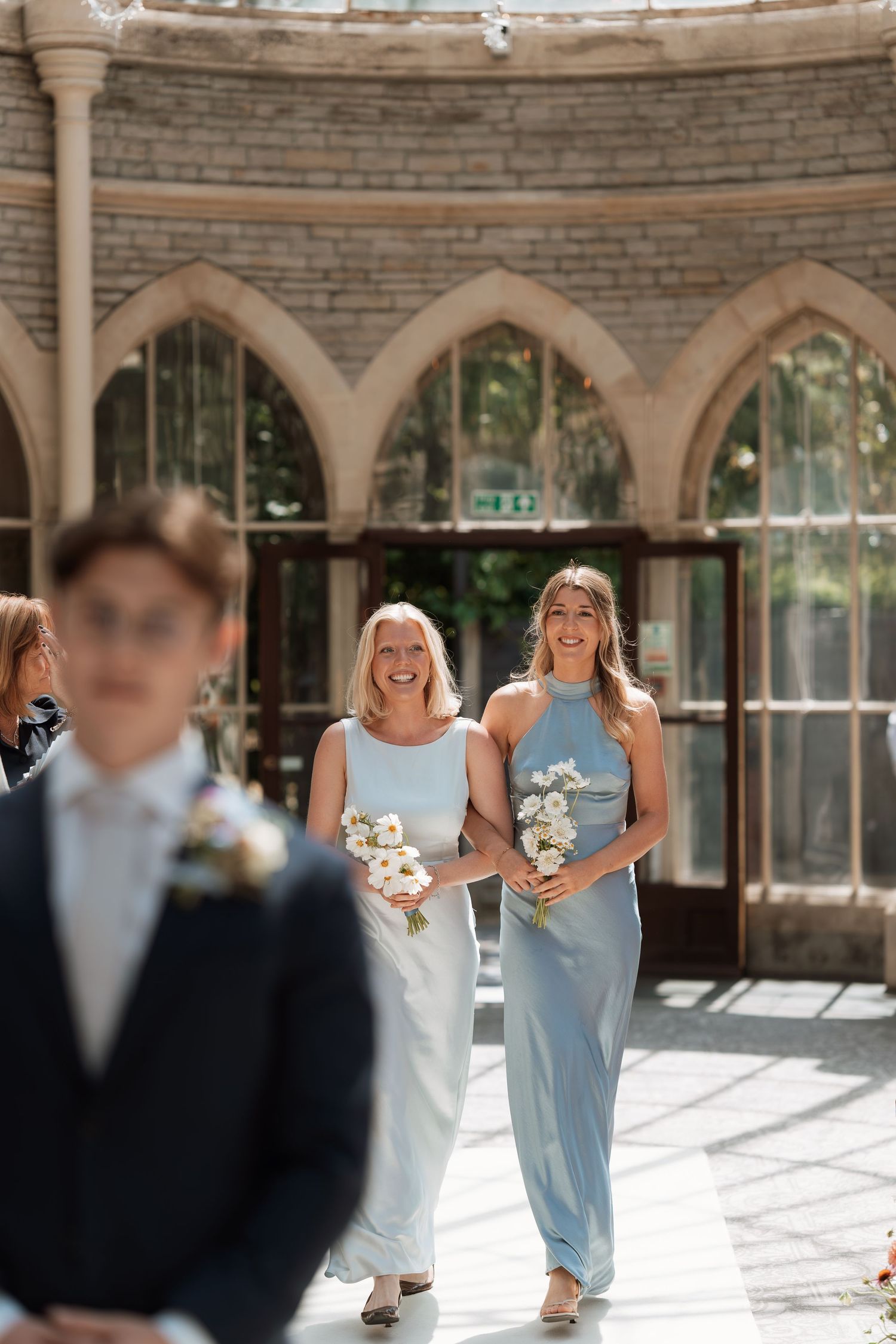 Bridesmaids in light blue dresses walk through a stone archway corridor filled with natural sunlight.
