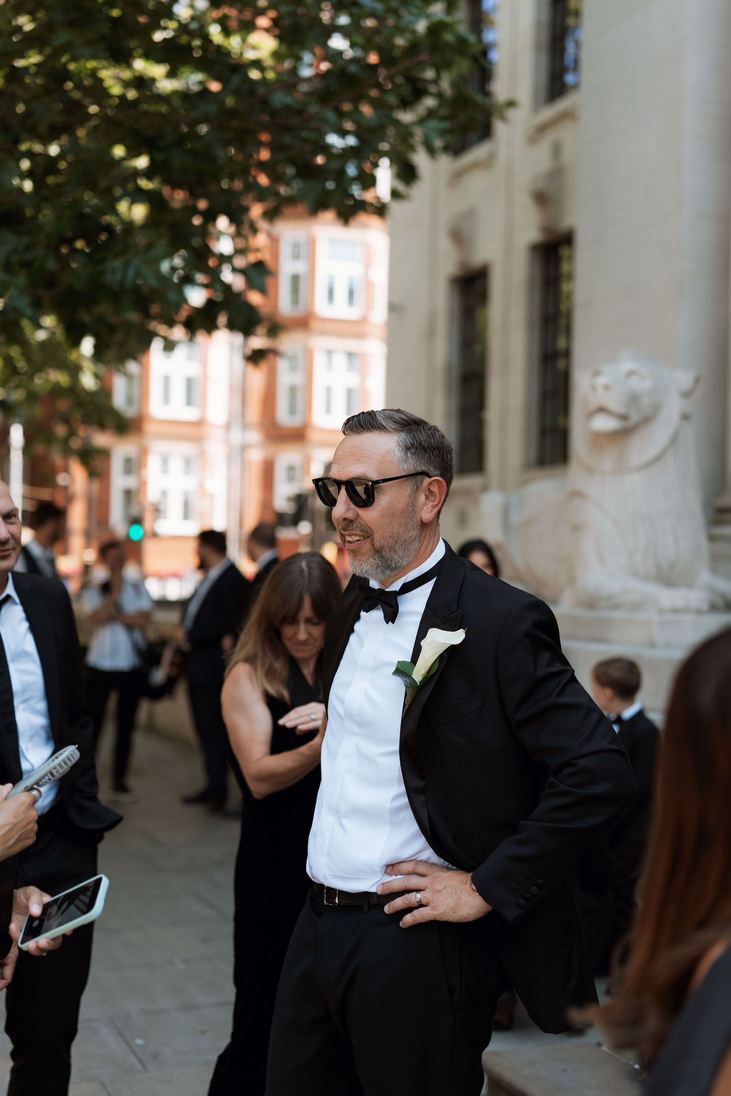Wedding attendee in a tuxedo and sunglasses stands outdoors near historic architecture.
