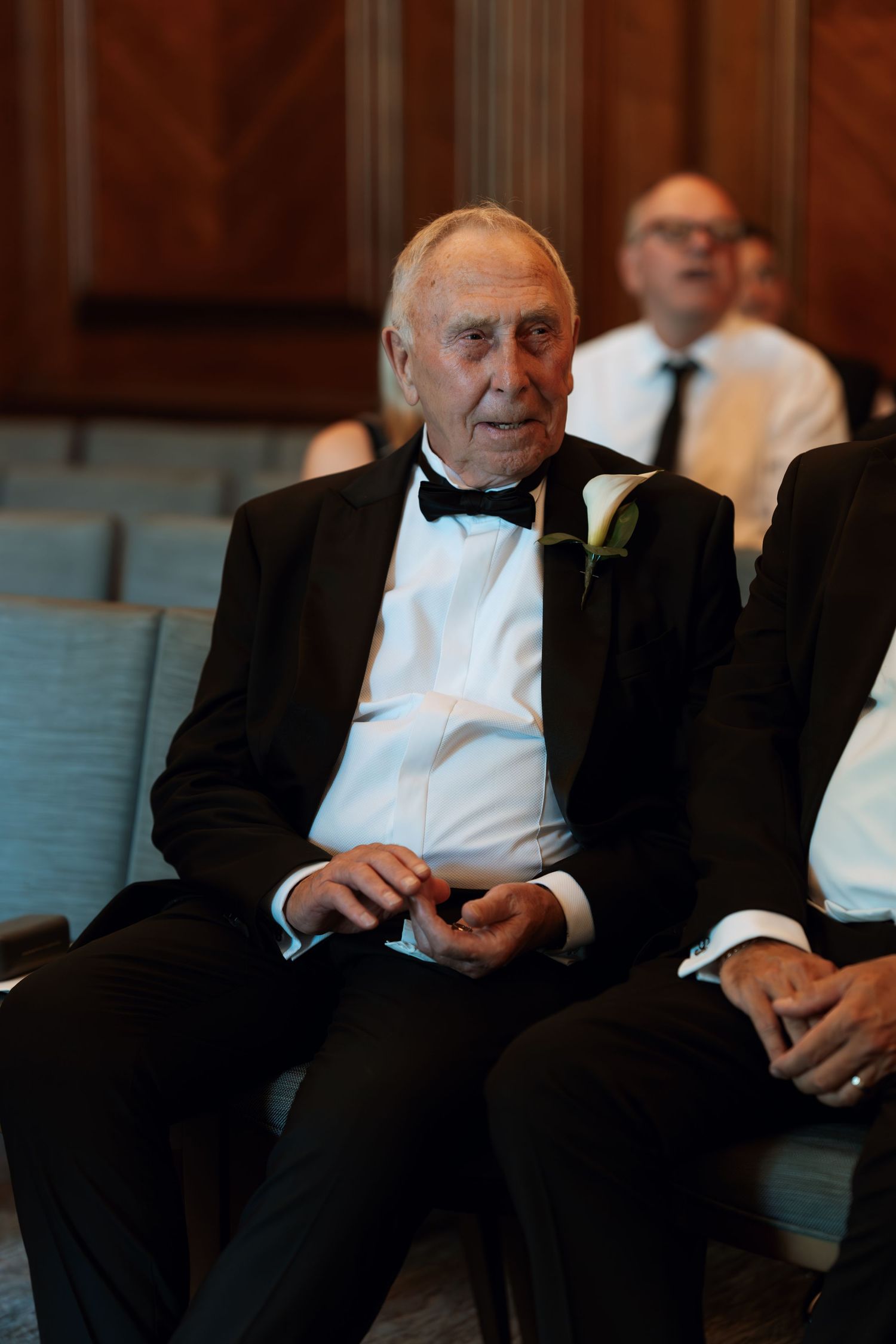 Two men in formal black tie attire sit in theater seats during an event.