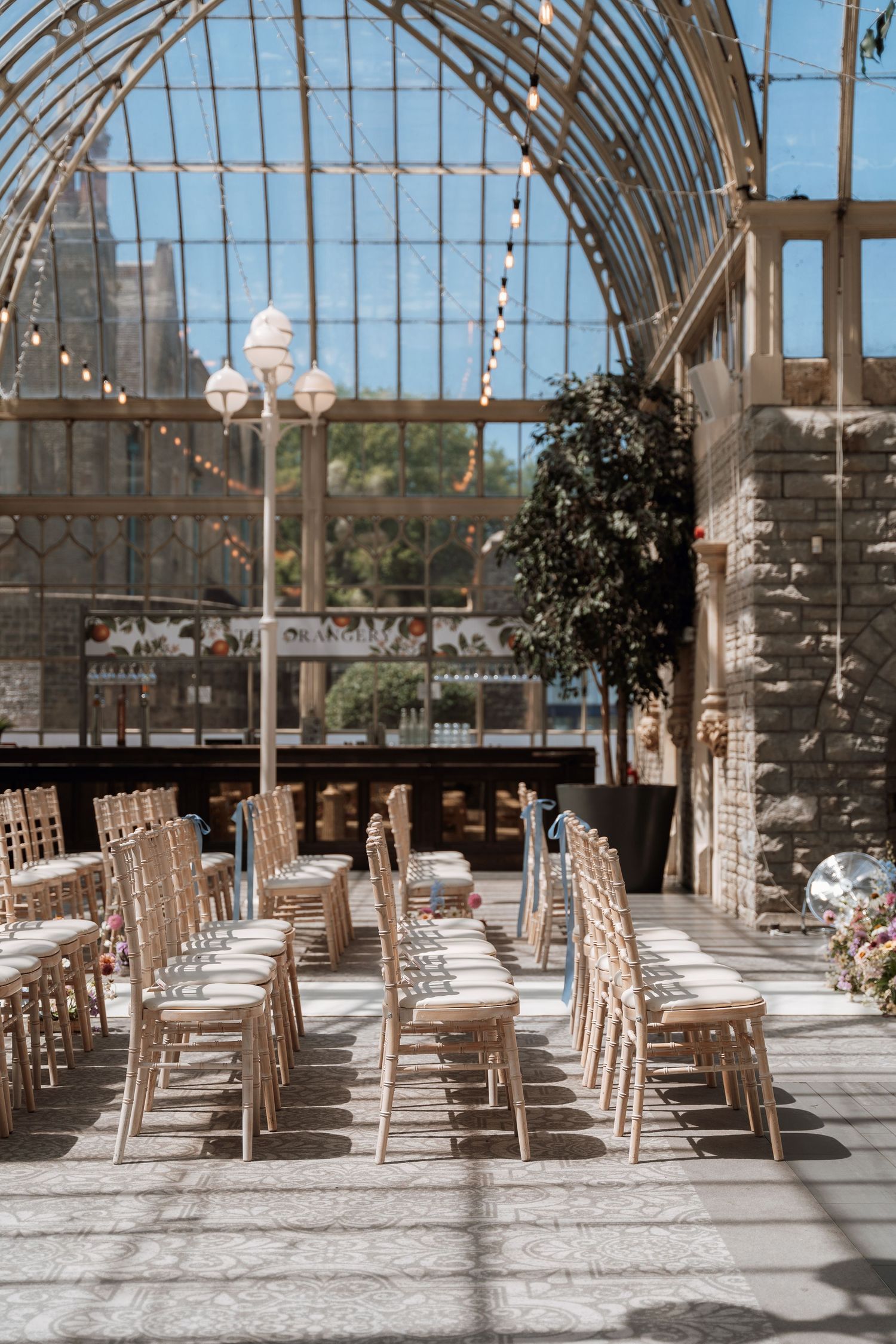 Light-filled conservatory event space with rows of wooden chairs set up for a wedding ceremony under a glass ceiling.