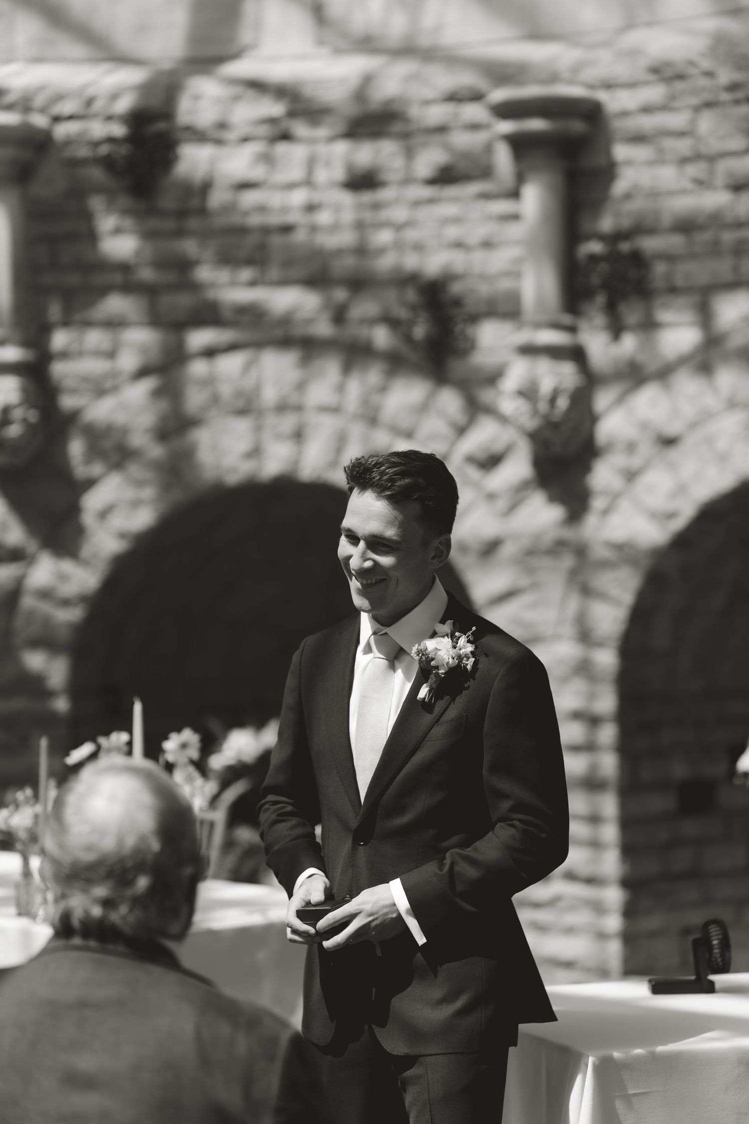 Black and white wedding photo of groom in suit smiling at outdoor venue with stone wall backdrop.