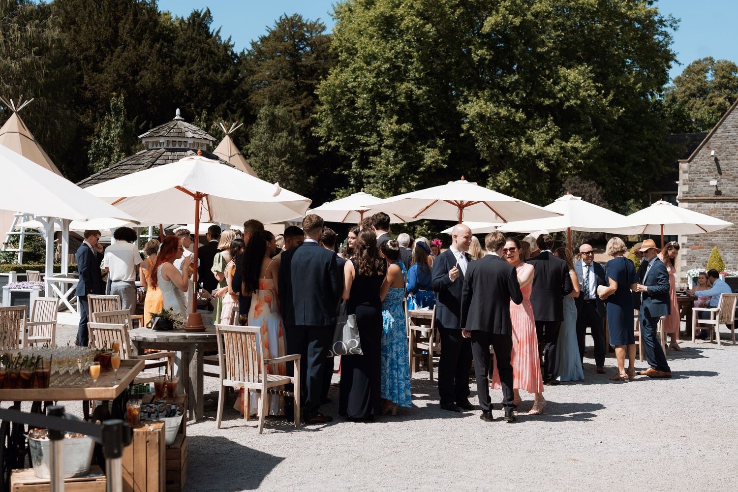 Outdoor event with white tents and people gathering at a sunny venue with trees in the background.