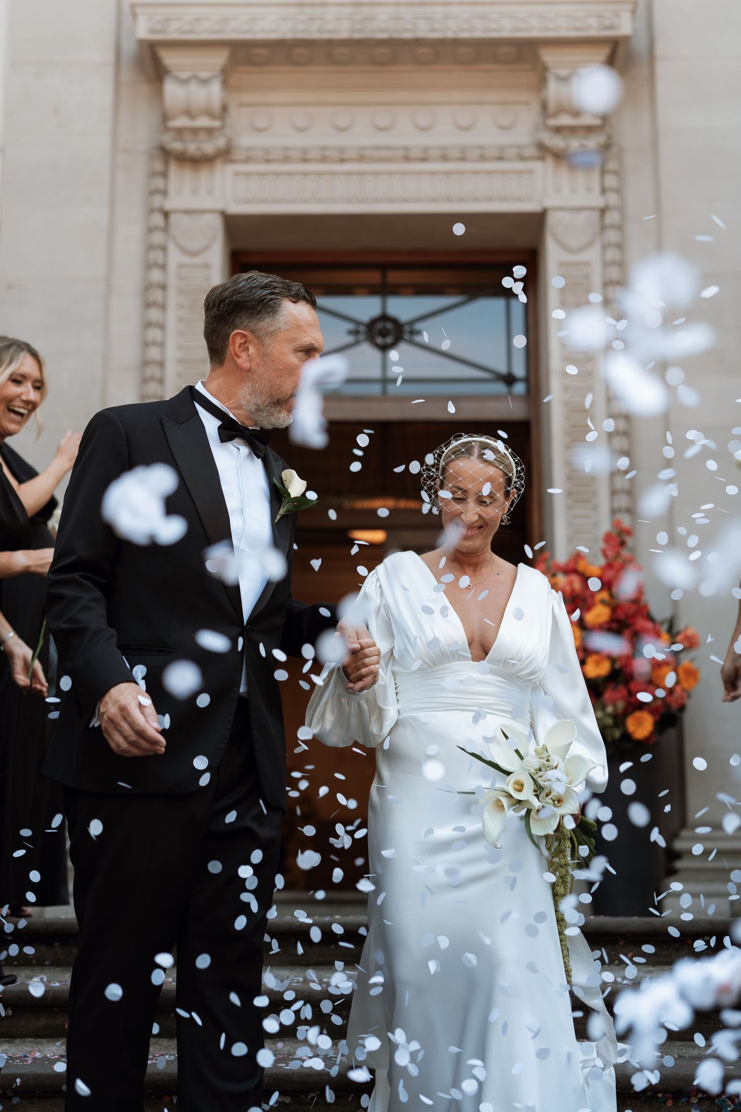 A couple exits a grand building entrance during their wedding celebration as confetti floats through the air.