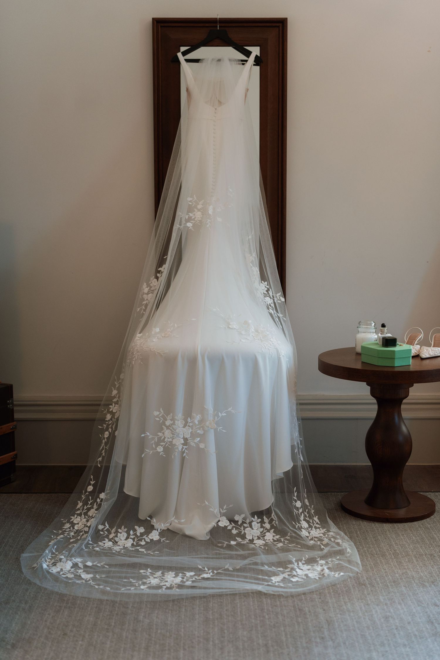 A white wedding dress with delicate floral lace veil hangs elegantly on a wooden wall while displayed on a hanger.