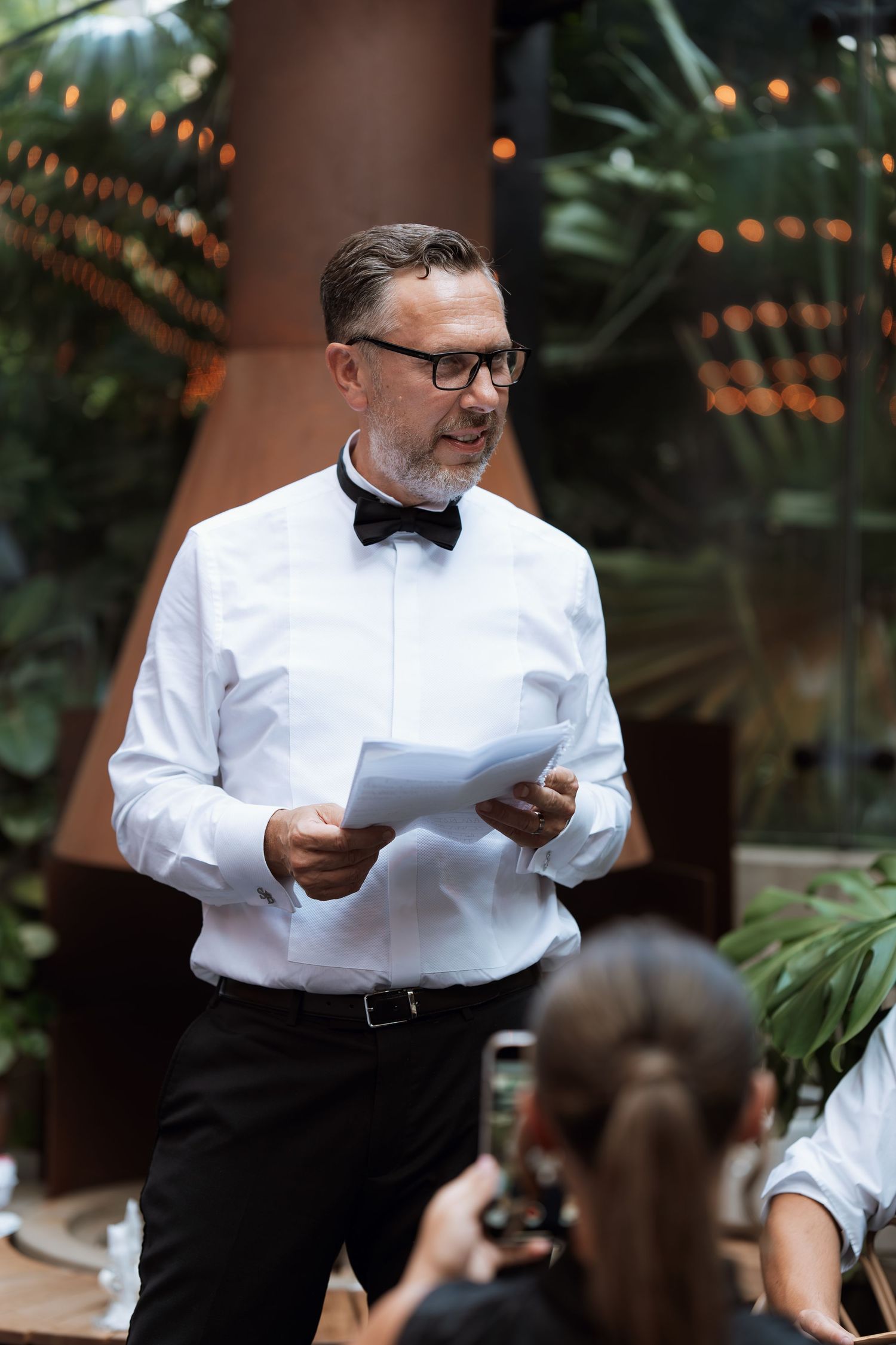 A person in formal attire giving a speech at an outdoor venue with string lights and tropical foliage.