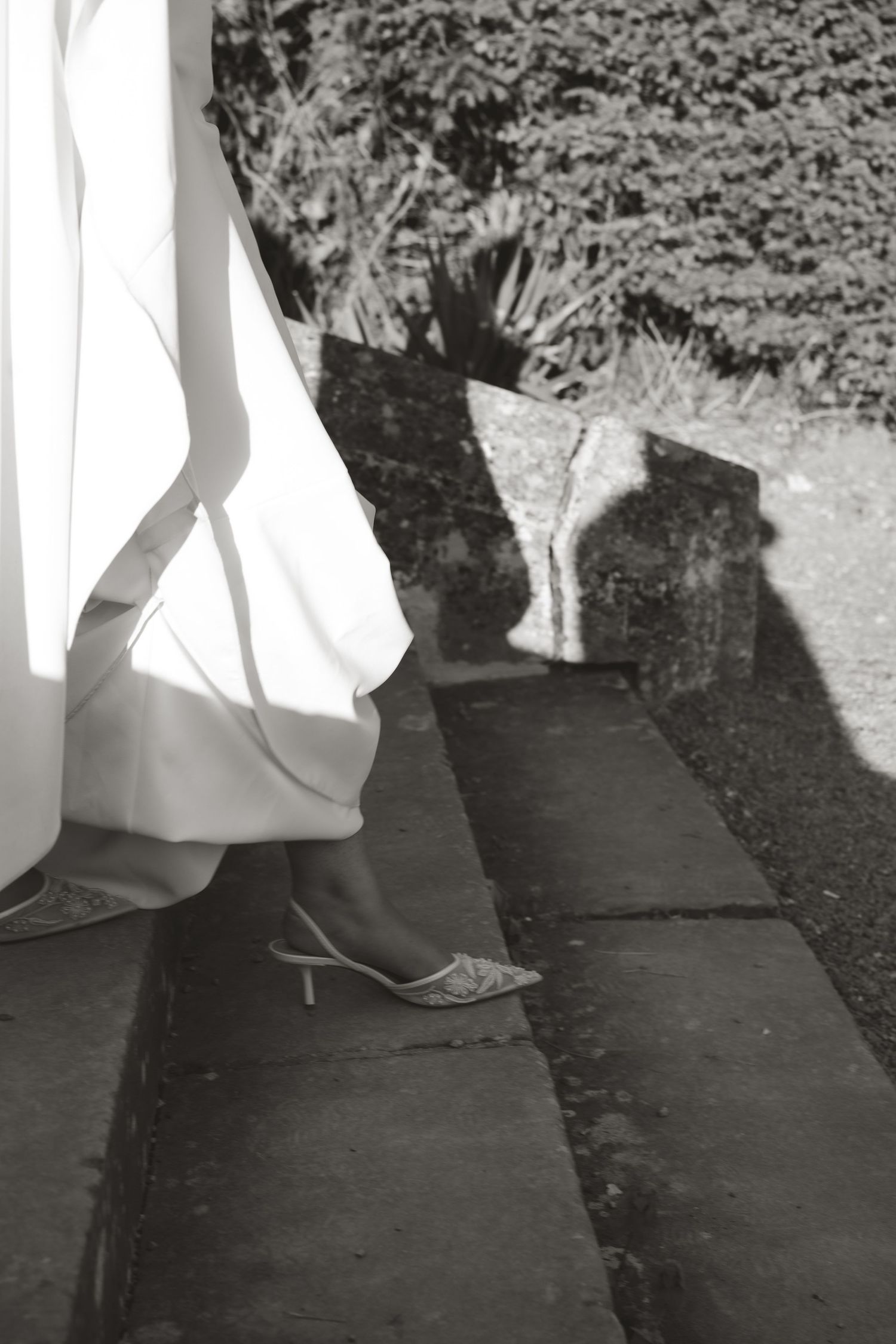 Black and white photo of a wedding dress hem and shoes casting shadows on stone steps.