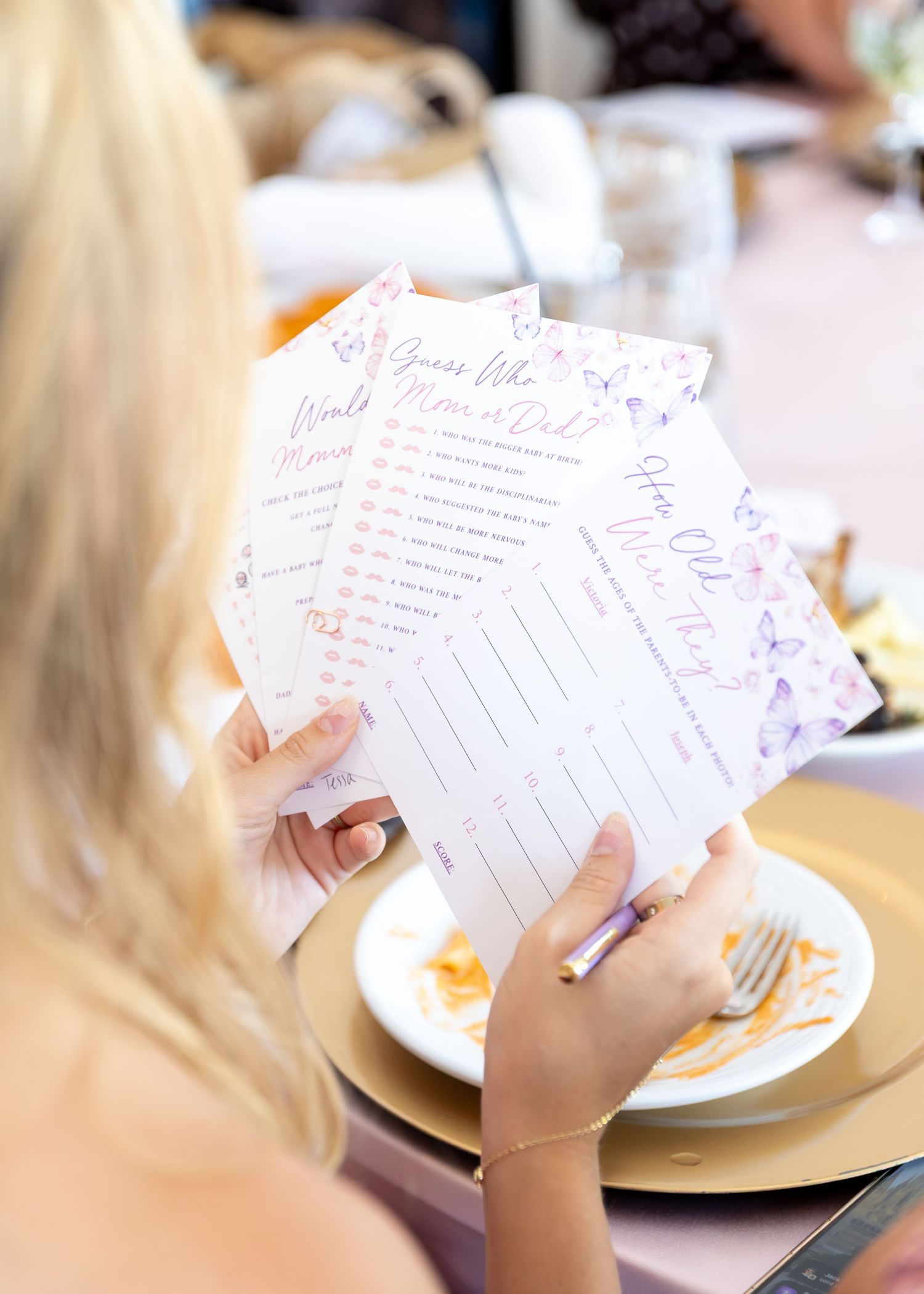 Guest holding baby shower game cards with butterfly design at a gold place setting during a baby shower.