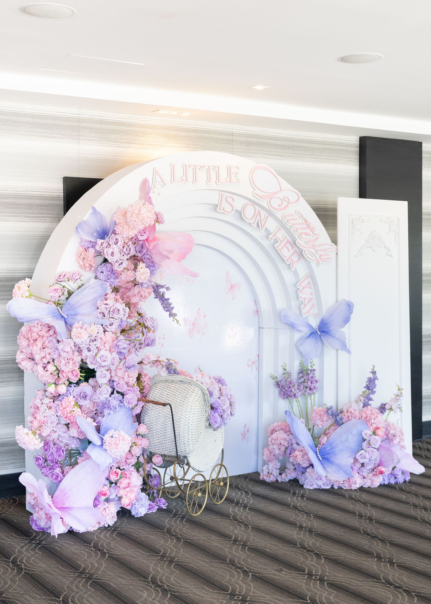 Stunning butterfly-themed baby shower backdrop with pink and purple floral arrangements and a vintage baby carriage.